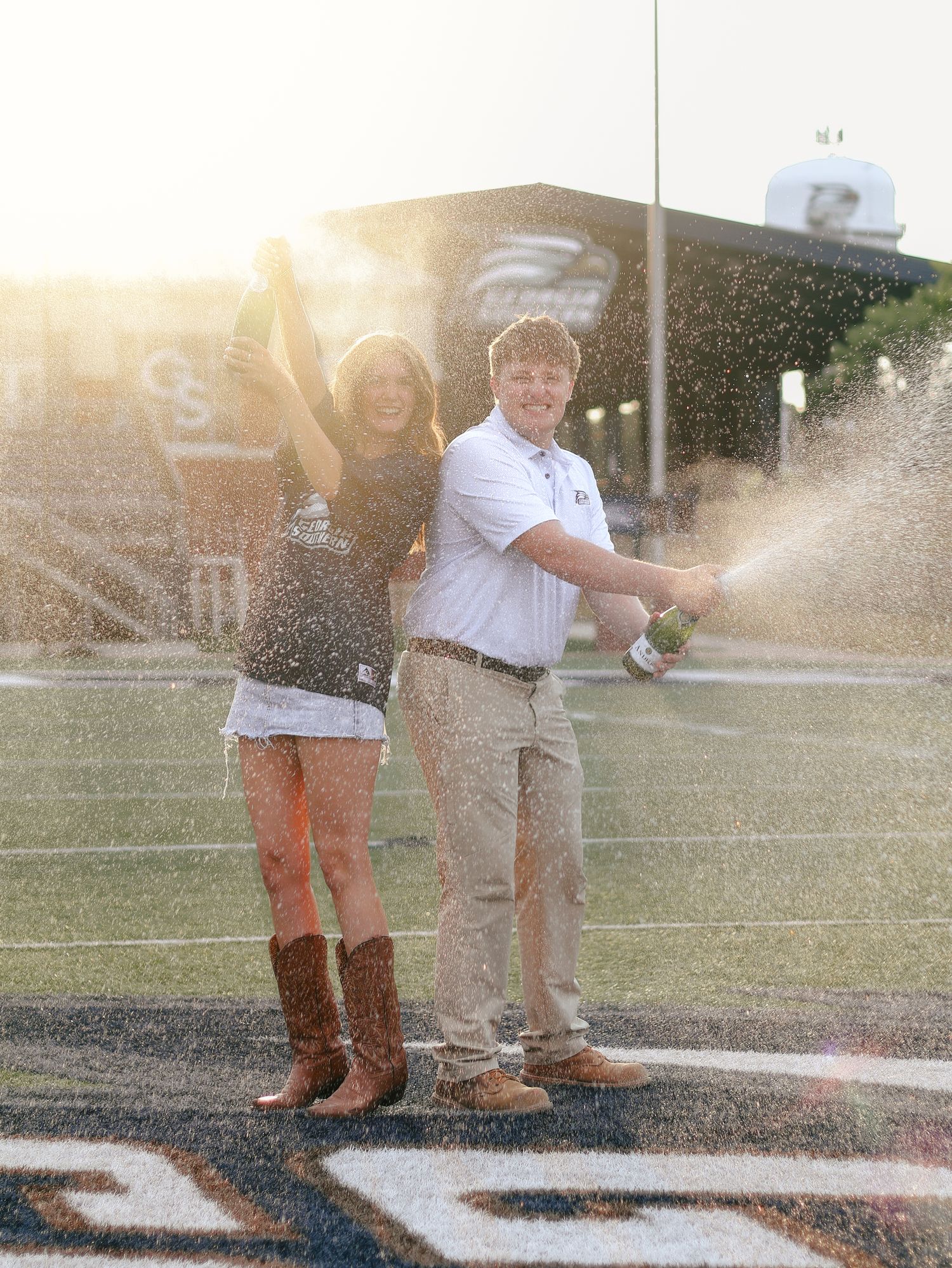 Couple in cowboy boots celebrates by spraying champagne on a football field at sunset, with stadium stands glowing in the background.