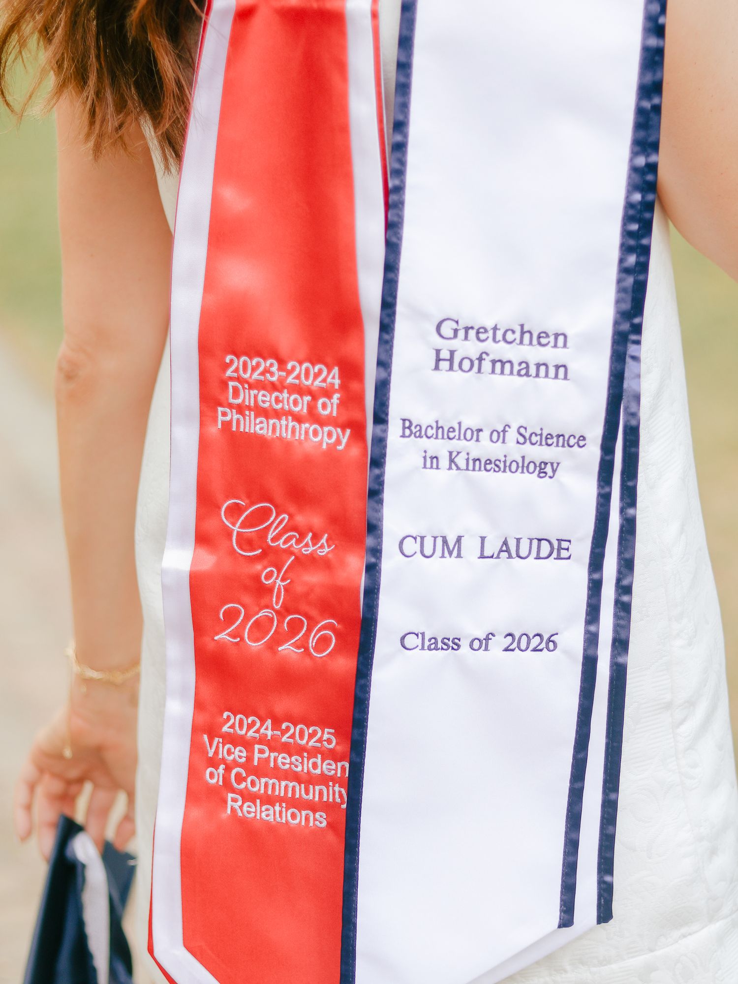 Graduate wearing a white and red graduation stole embroidered with Gretchen Hofmann, Kinesiology degree, cum laude, Class of 2026.
