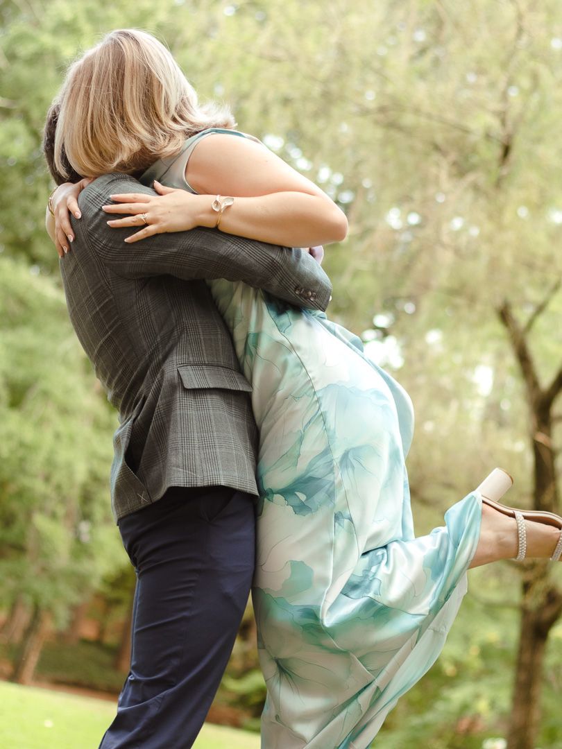 A man in a gray plaid blazer lifts a woman in a flowing teal dress in a joyful embrace among green trees.