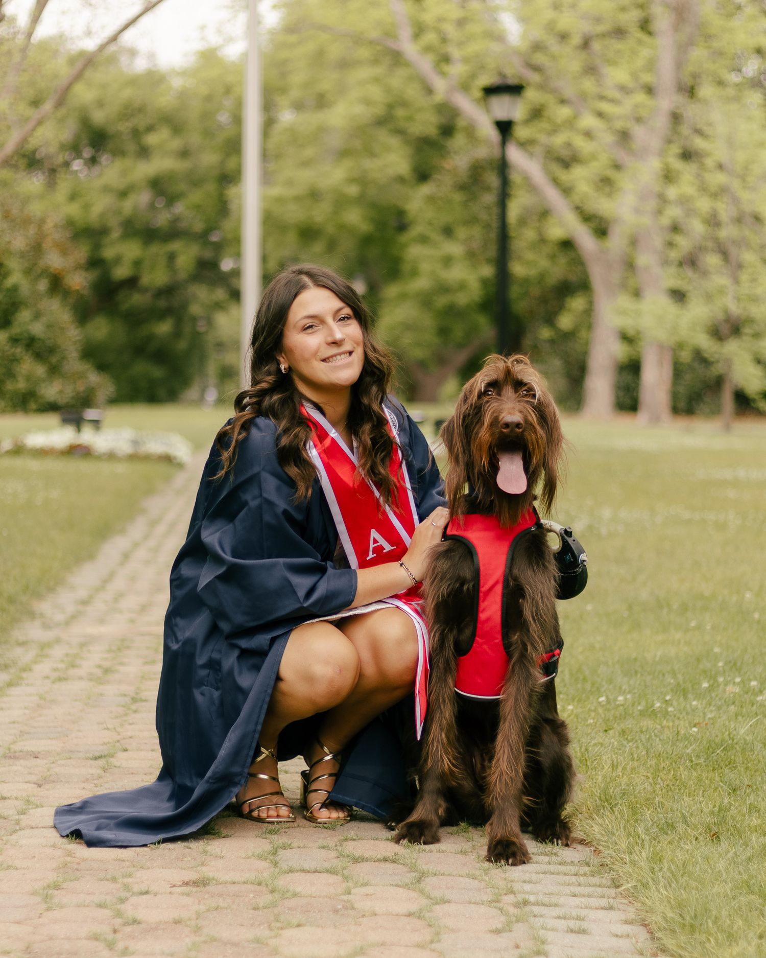 A smiling graduate in a navy gown and red sash kneels on a brick path beside her happy brown dog wearing a matching red vest.