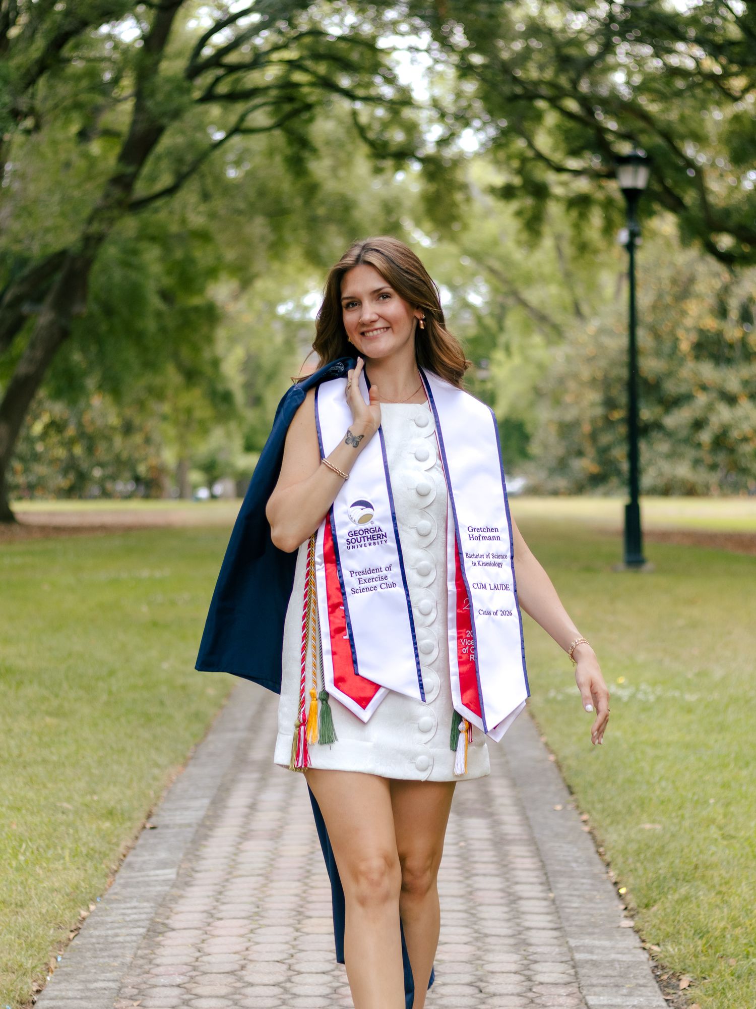 Smiling college graduate in a white dress and graduation stole holding a navy gown while walking along a brick path under shady trees.