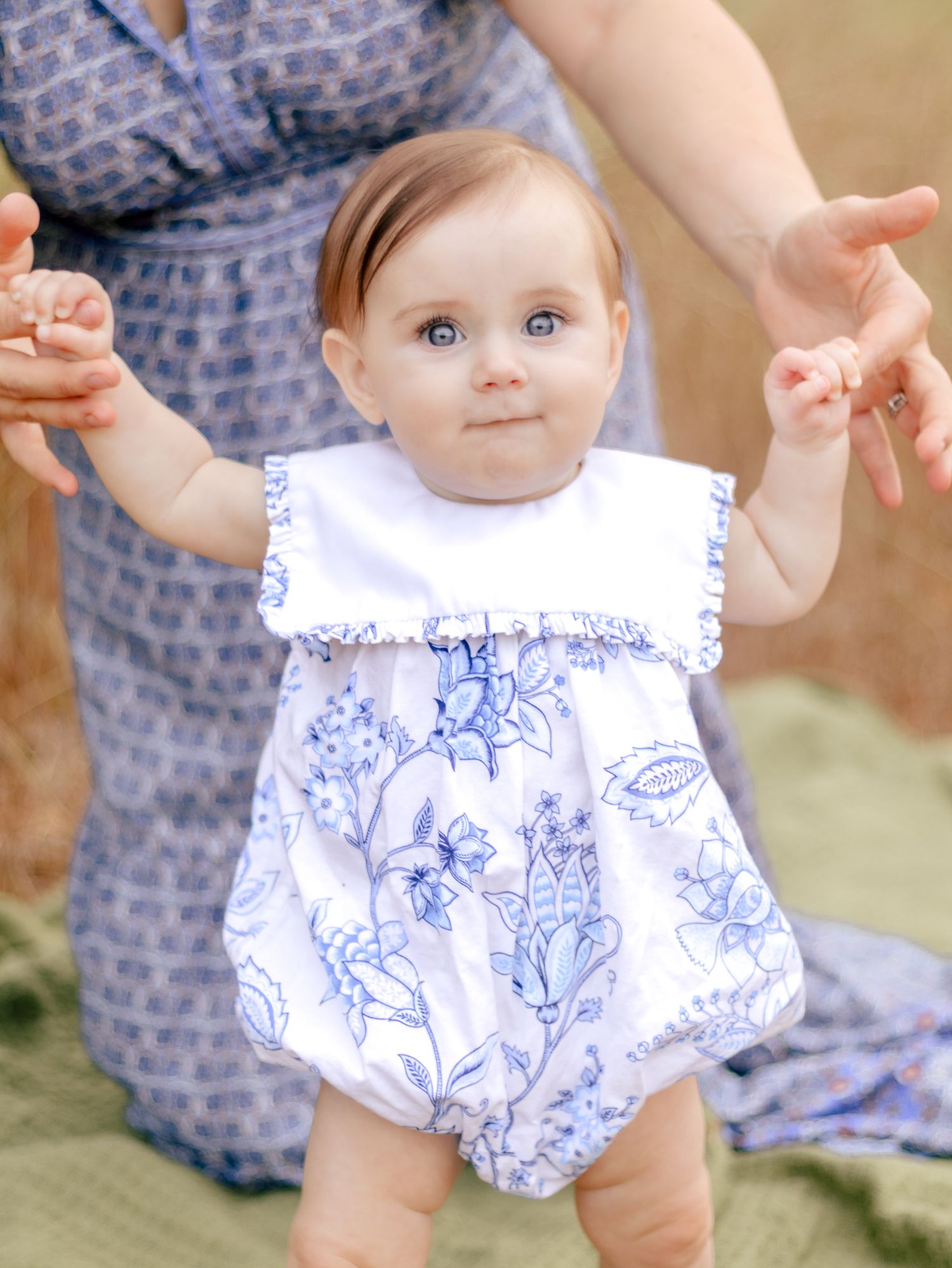 Baby in a white and blue floral bubble romper with a ruffled bib collar stands on a green blanket while a parent holds her hands.