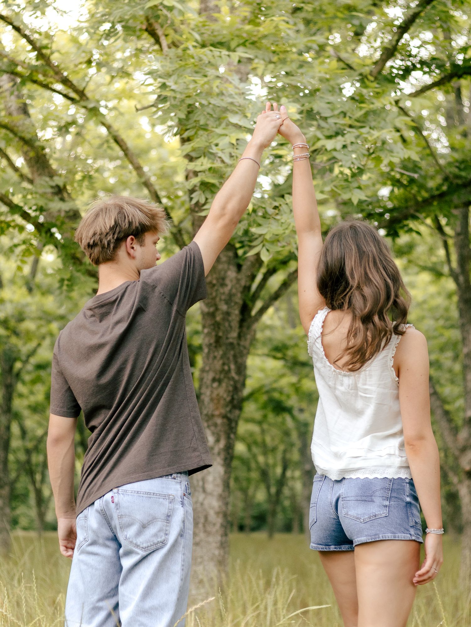 Couple seen from behind holding raised hands together while standing in a sunlit grassy meadow surrounded by green leafy trees.