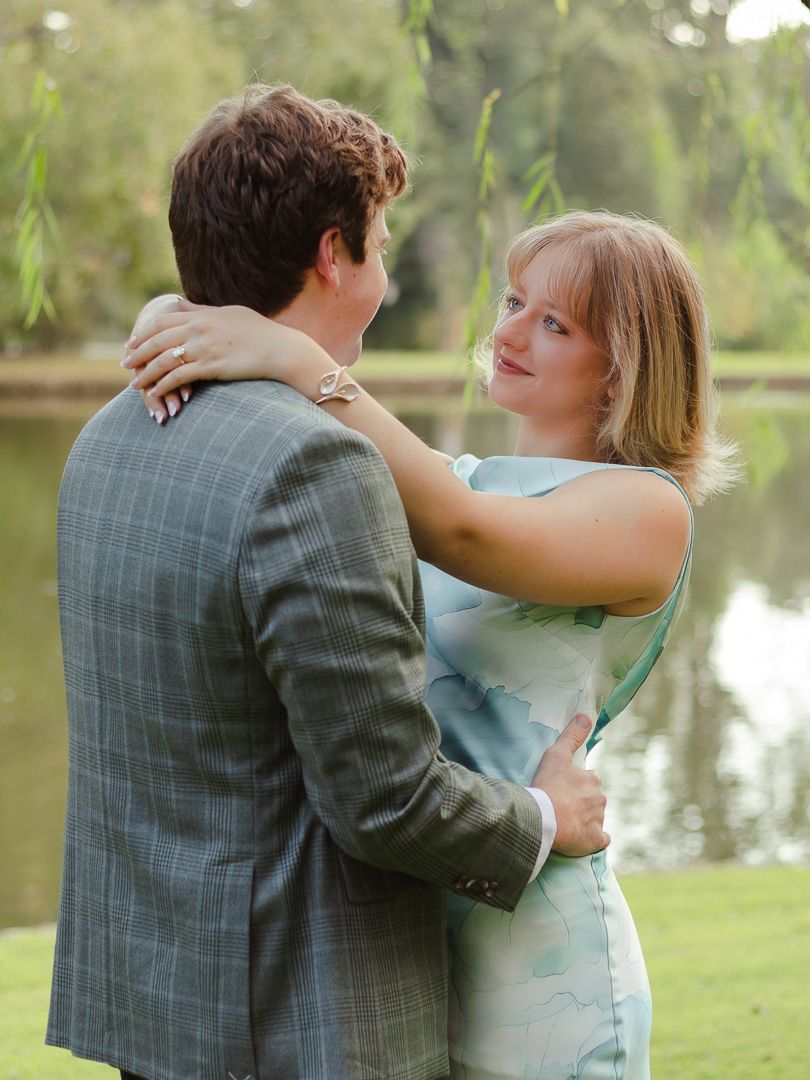Engaged couple embracing by a calm lake, the woman in a light blue dress with her arm around the man in a plaid blazer.