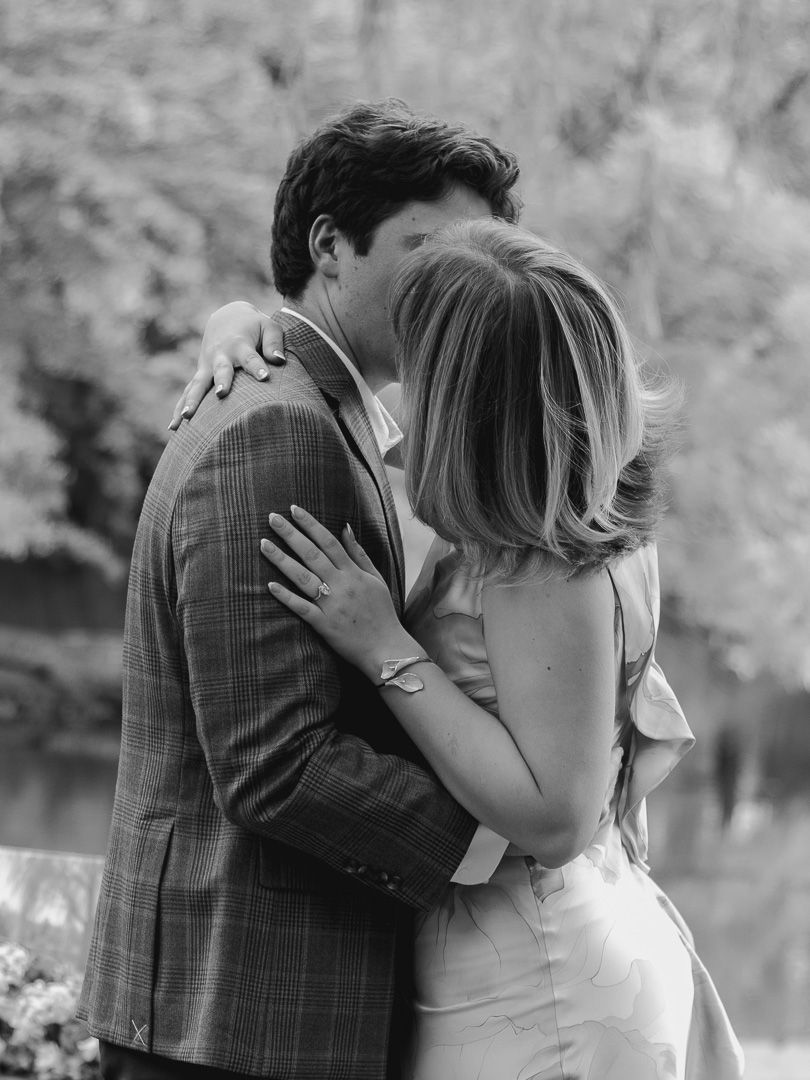 Black and white photo of an engaged couple kissing by the water, the woman's ring and bracelet visible as her hand rests on his chest.