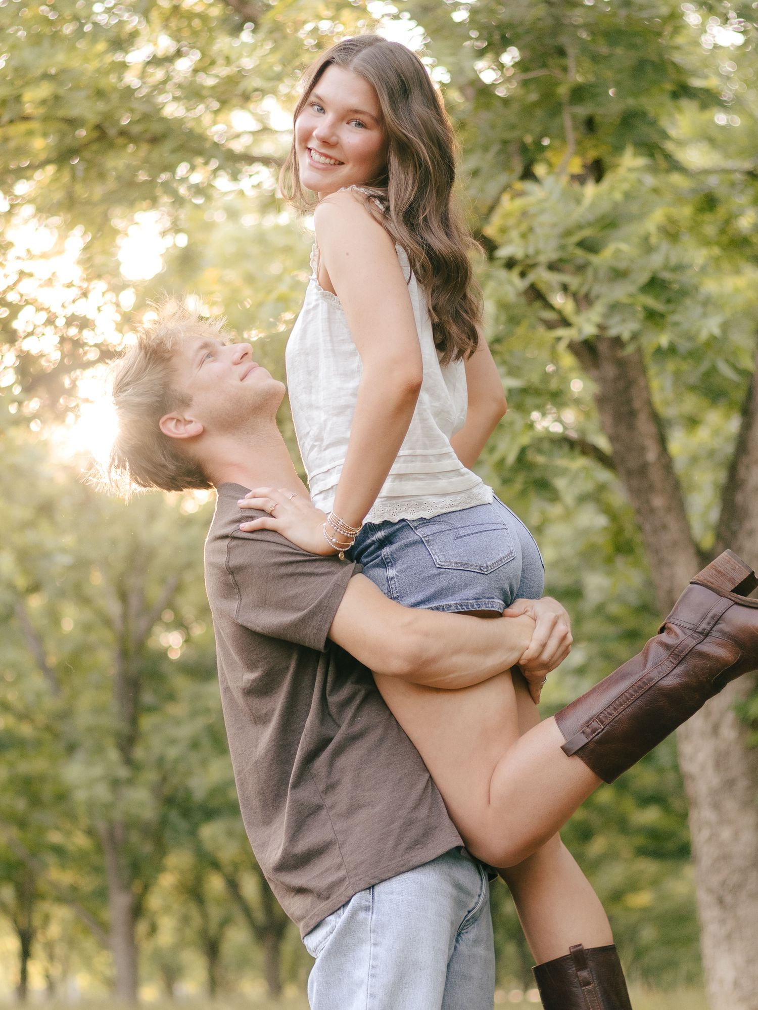 A smiling couple in casual clothes and brown boots pose outdoors as a man lifts a woman in the air among sunlit trees.