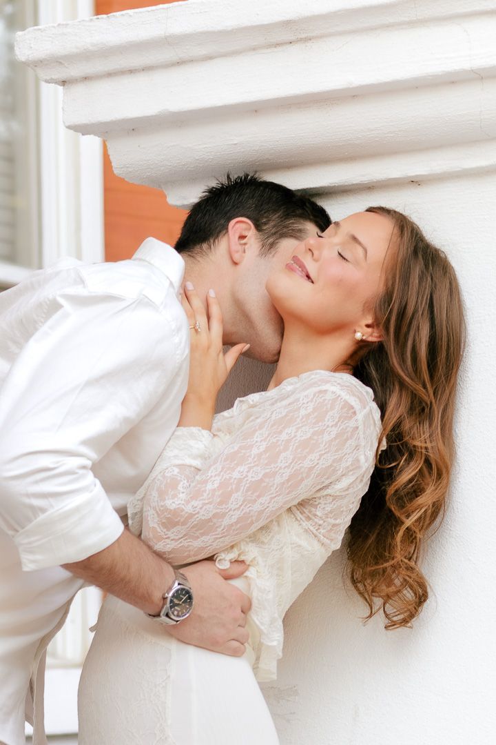 Romantic couple embracing against a white wall as a smiling woman in a lace blouse closes her eyes while a man kisses her neck.