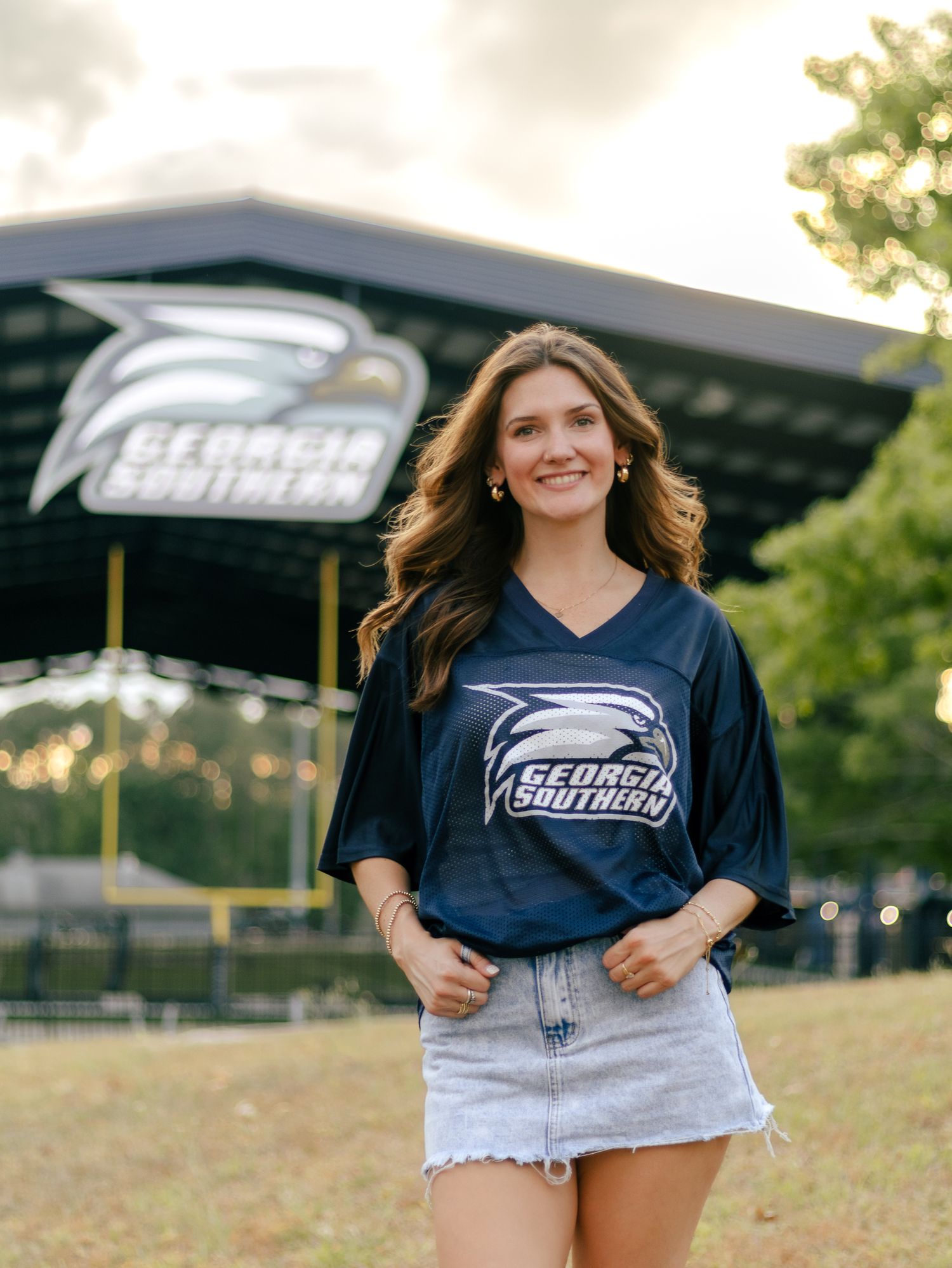 Smiling young woman in a navy Georgia Southern jersey and denim skirt stands on grass in front of a Georgia Southern eagle logo stadium.