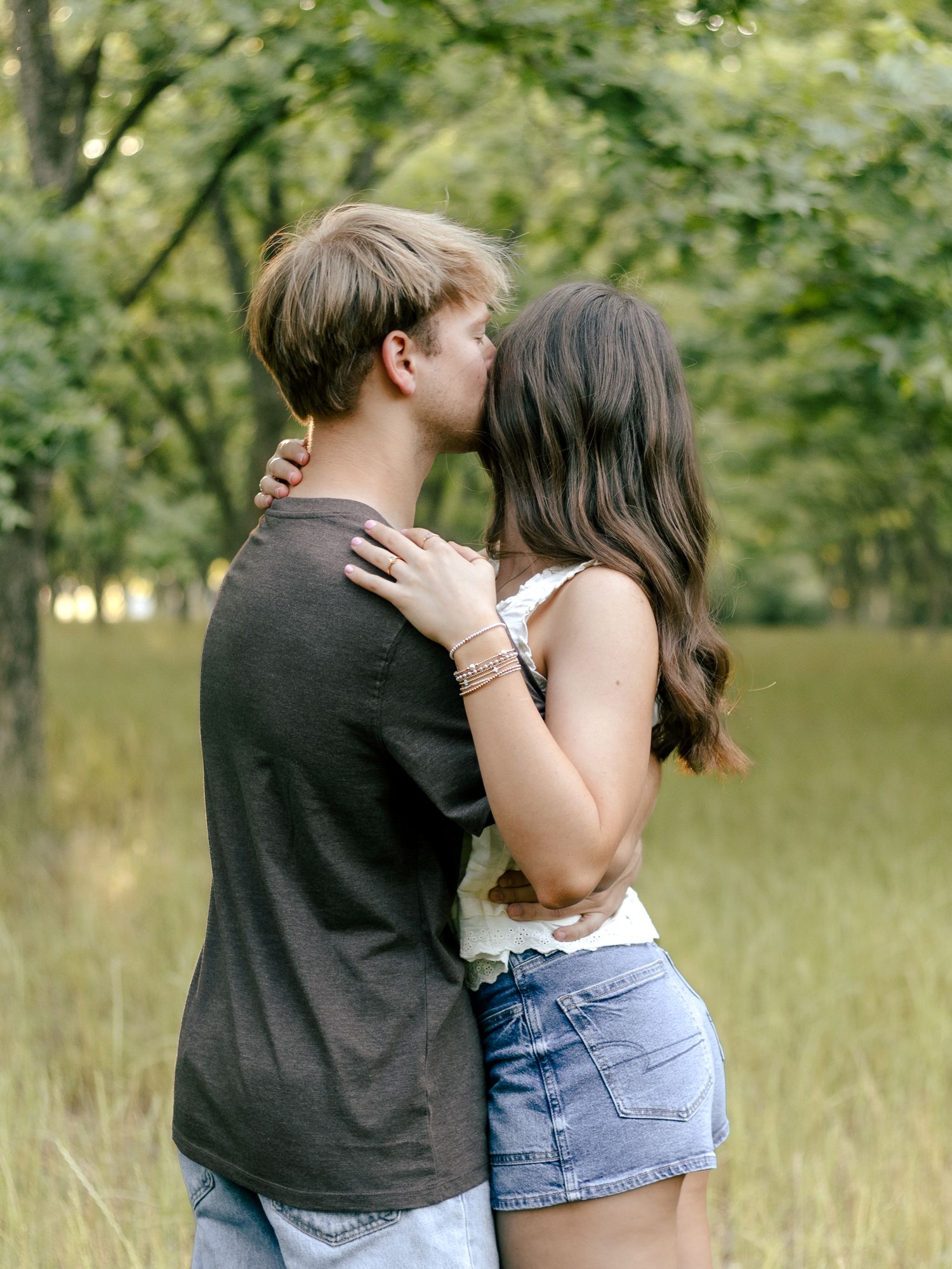 A young couple embraces in a sunlit grassy field surrounded by green trees as he gently kisses her forehead.