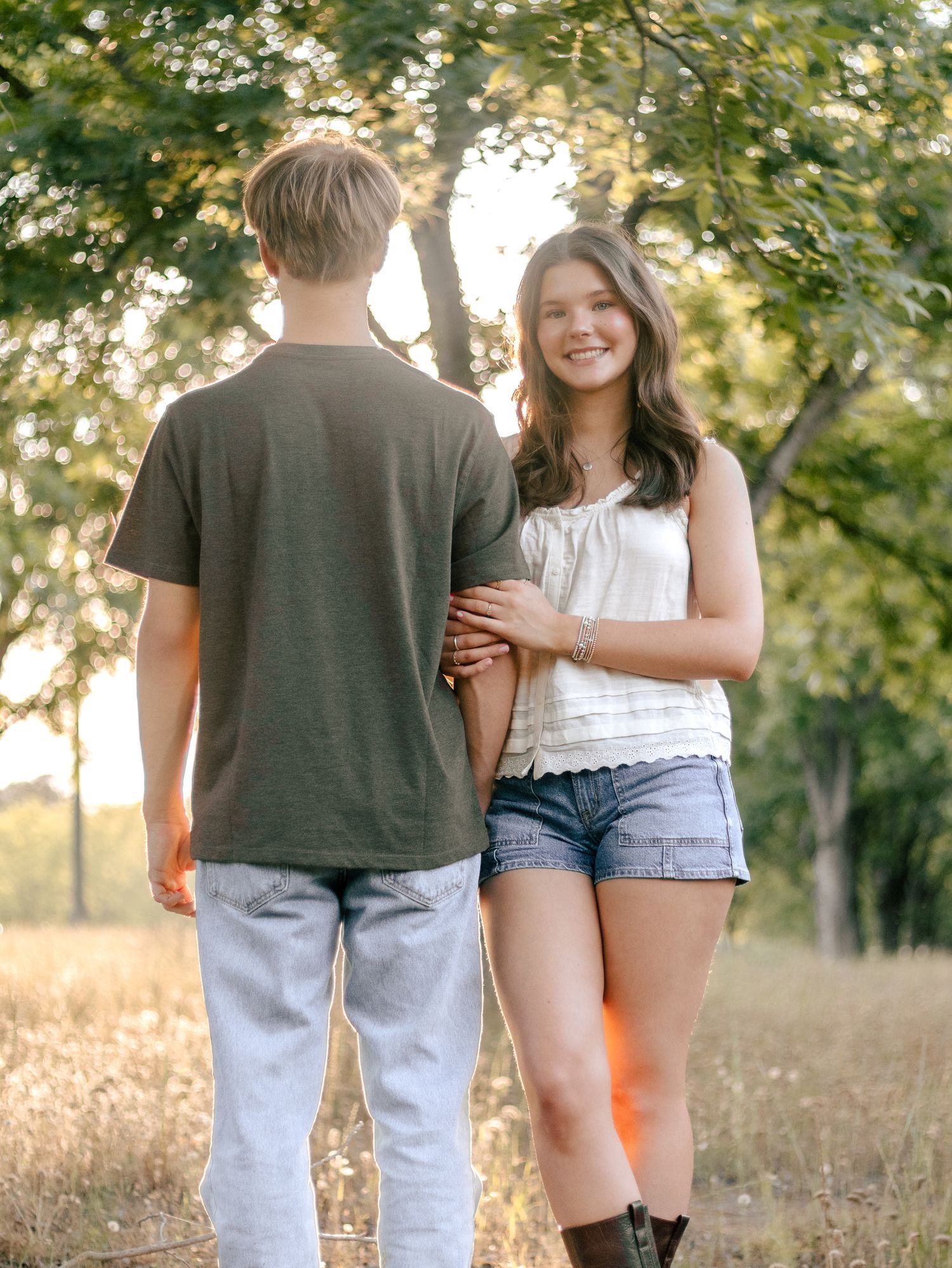 Smiling young woman in a white top, denim shorts and boots holds the arm of a young man facing away in a green tee in a sunlit grassy field.
