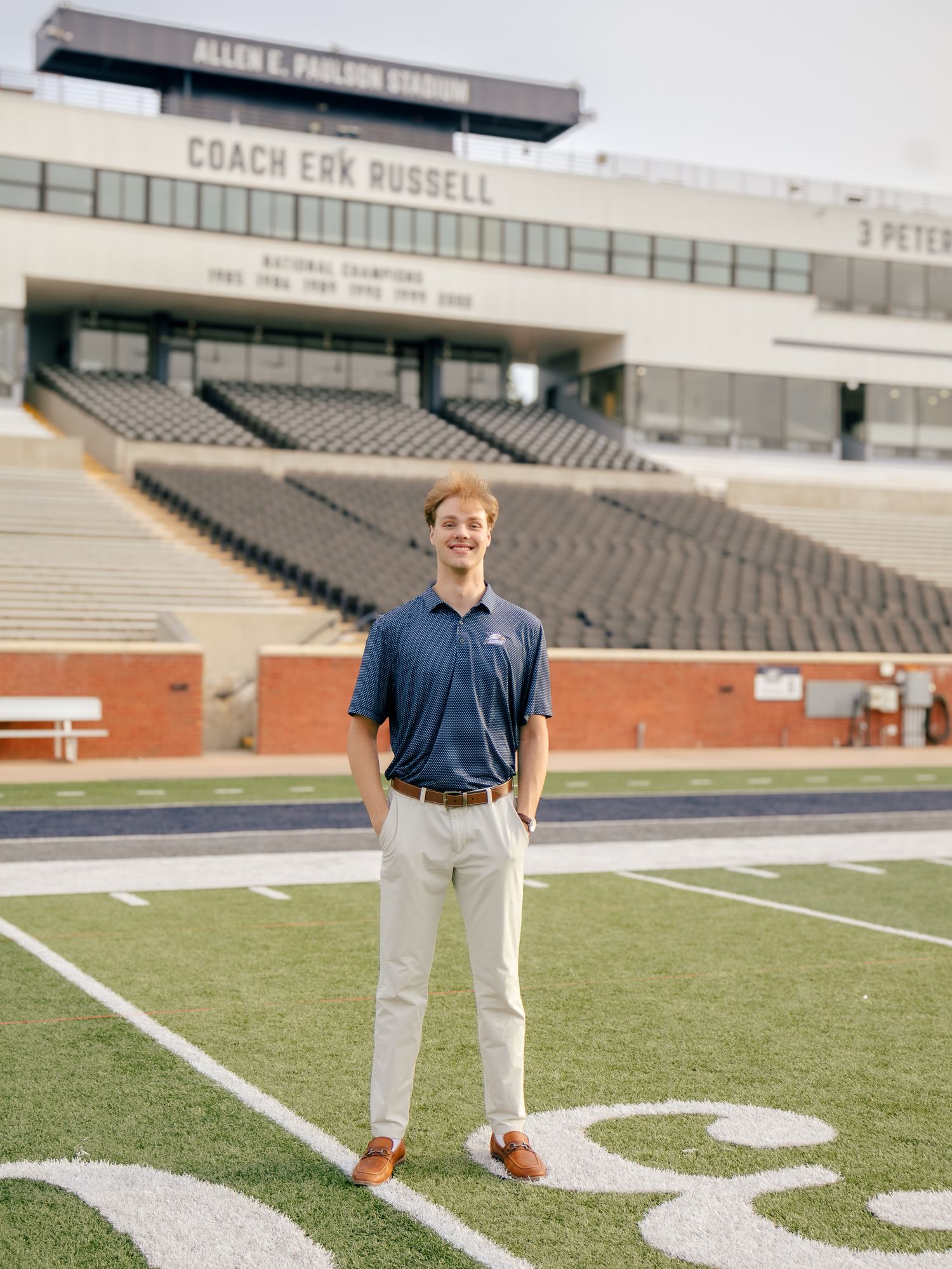 Young man in a navy polo, khaki pants and brown shoes stands on the turf of Allen E. Paulson Stadium with empty stands behind him.