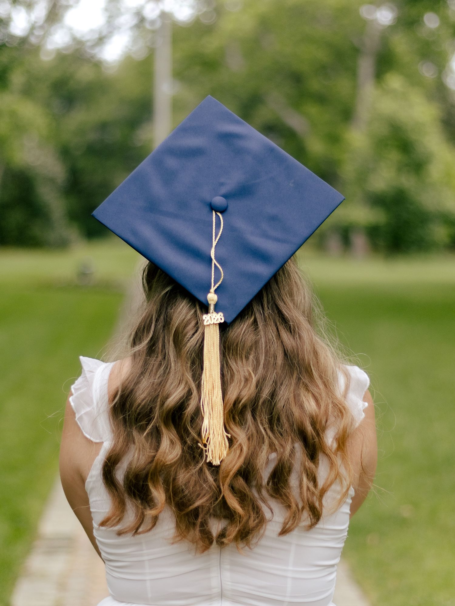Graduate seen from behind wearing a navy blue cap with a gold tassel and a white dress, standing on a path in a green park.