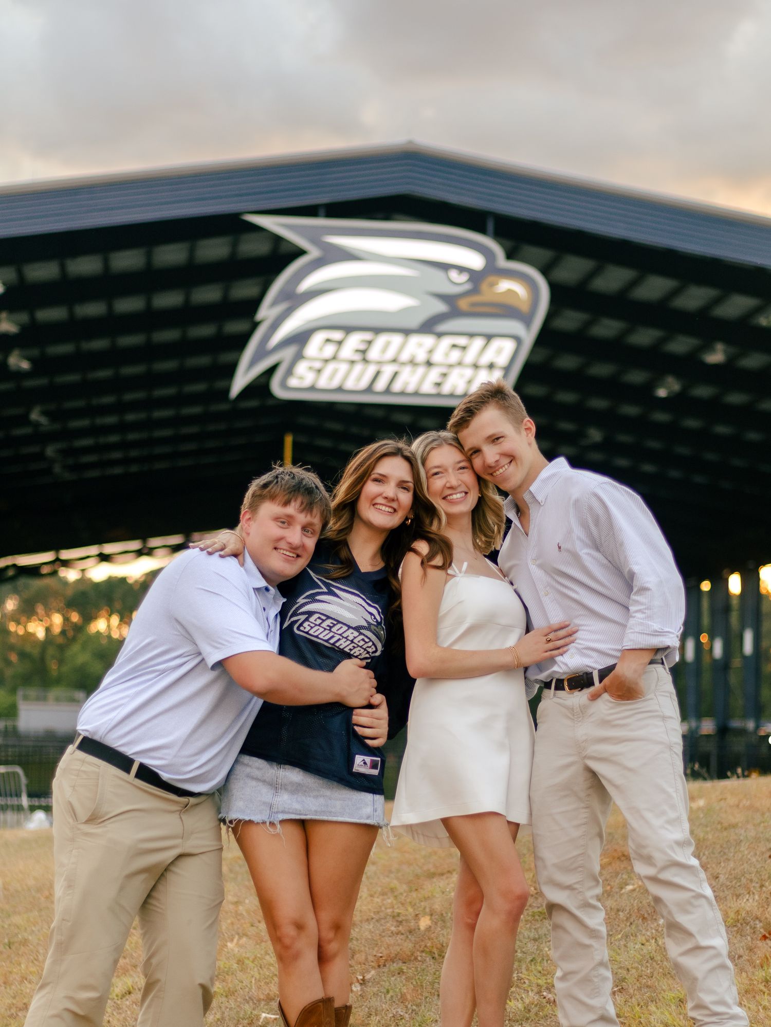 Four friends smile and hug in front of the Georgia Southern eagle logo on a stadium canopy at sunset.