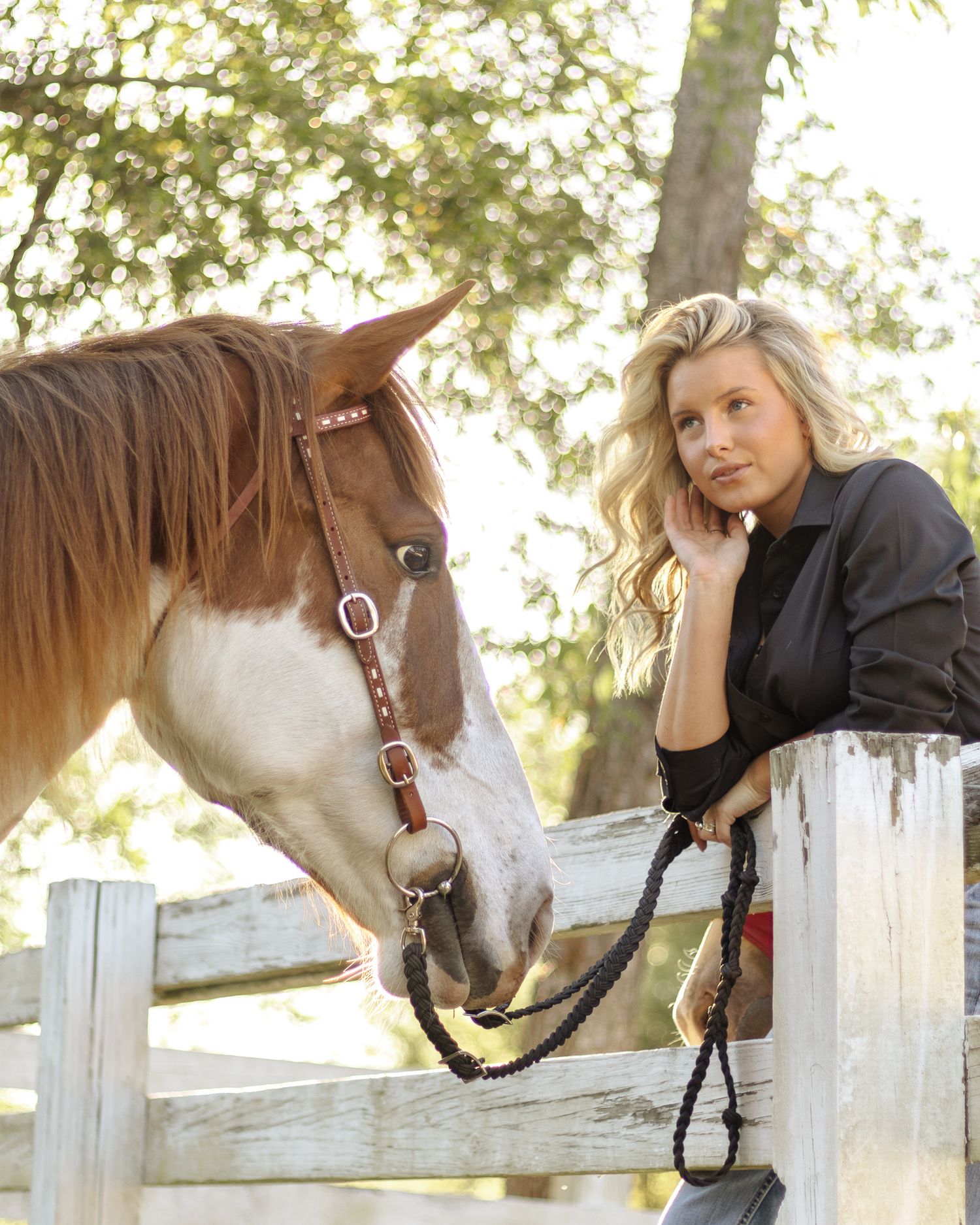 Blonde woman in a dark shirt rests her hand on her cheek beside a brown and white paint horse at a white wooden fence.