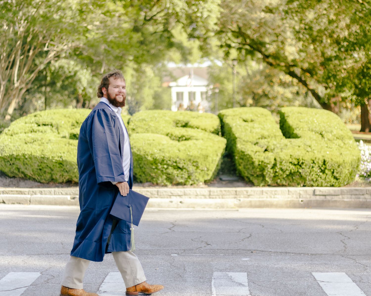Bearded man in a long blue robe carrying a book walks across a crosswalk past trimmed green hedges with a white building behind.