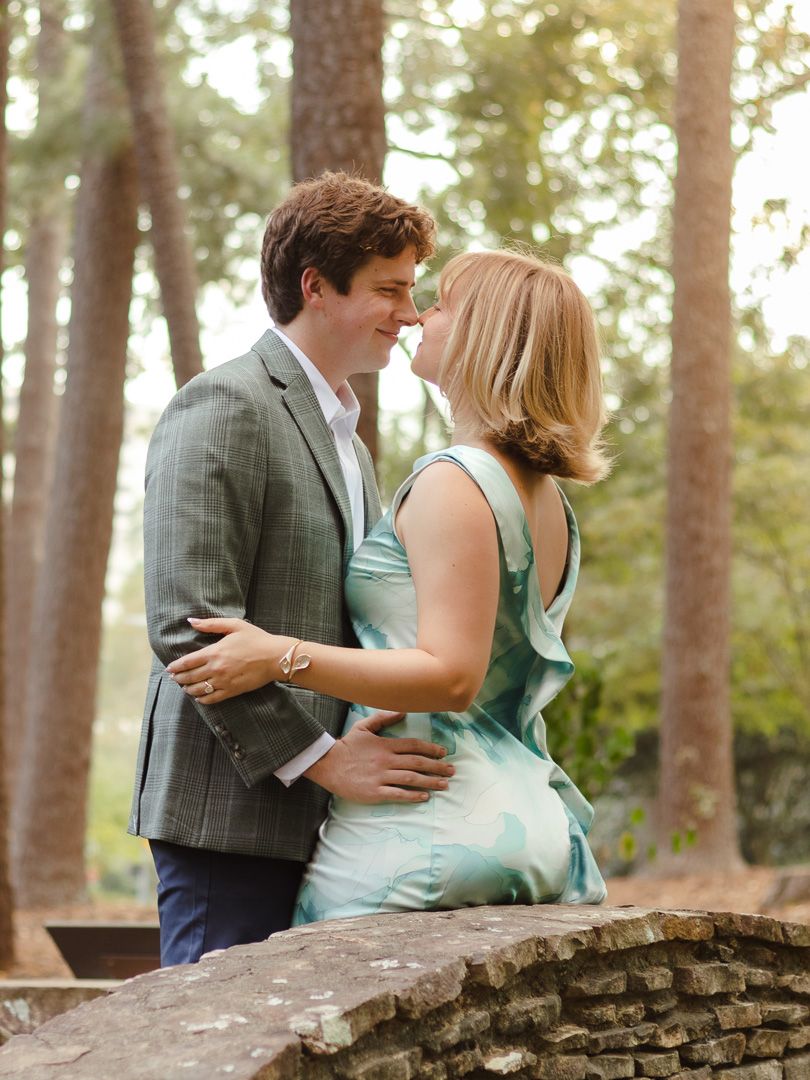 A couple in a gray plaid blazer and teal floral dress share an intimate moment on a stone wall among tall pine trees.