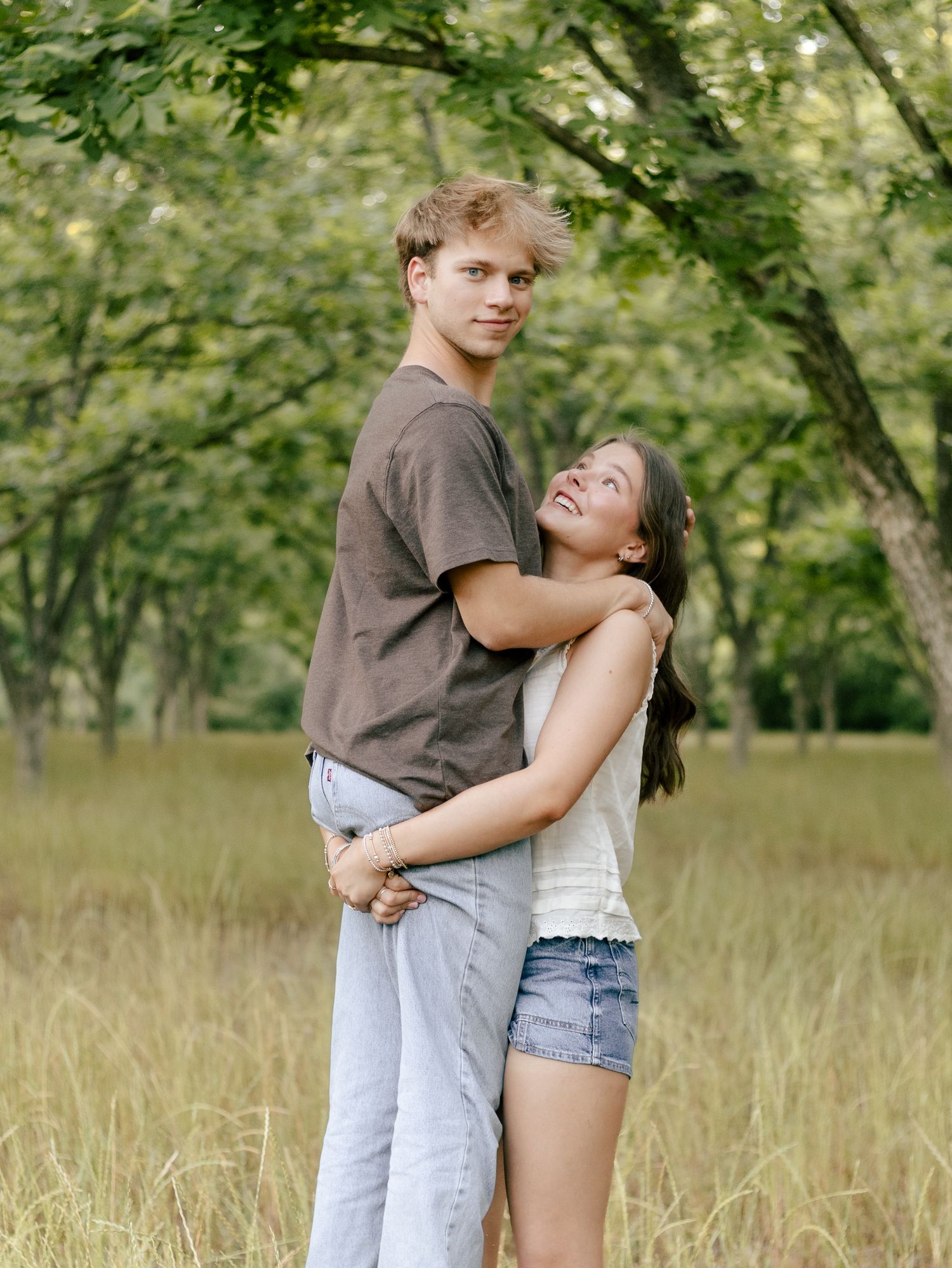 A young couple embraces in a sunlit grassy field surrounded by trees, the girl smiling up at the tall boy.