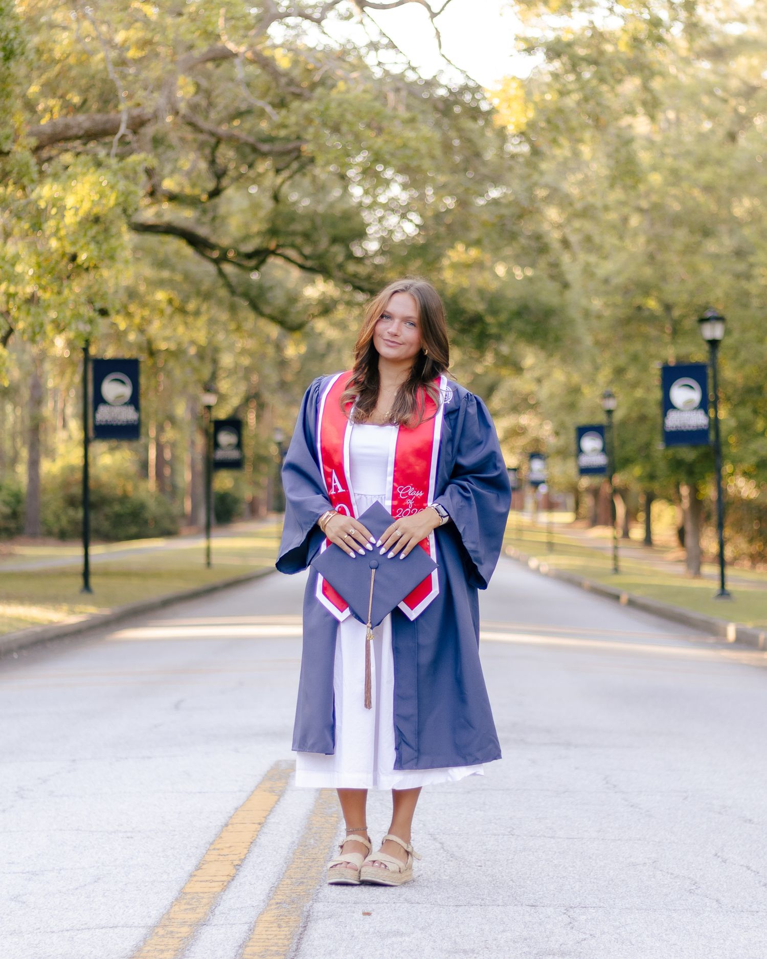 A college graduate in a navy gown and red stole holds her cap while standing on a tree-lined campus road with banners.