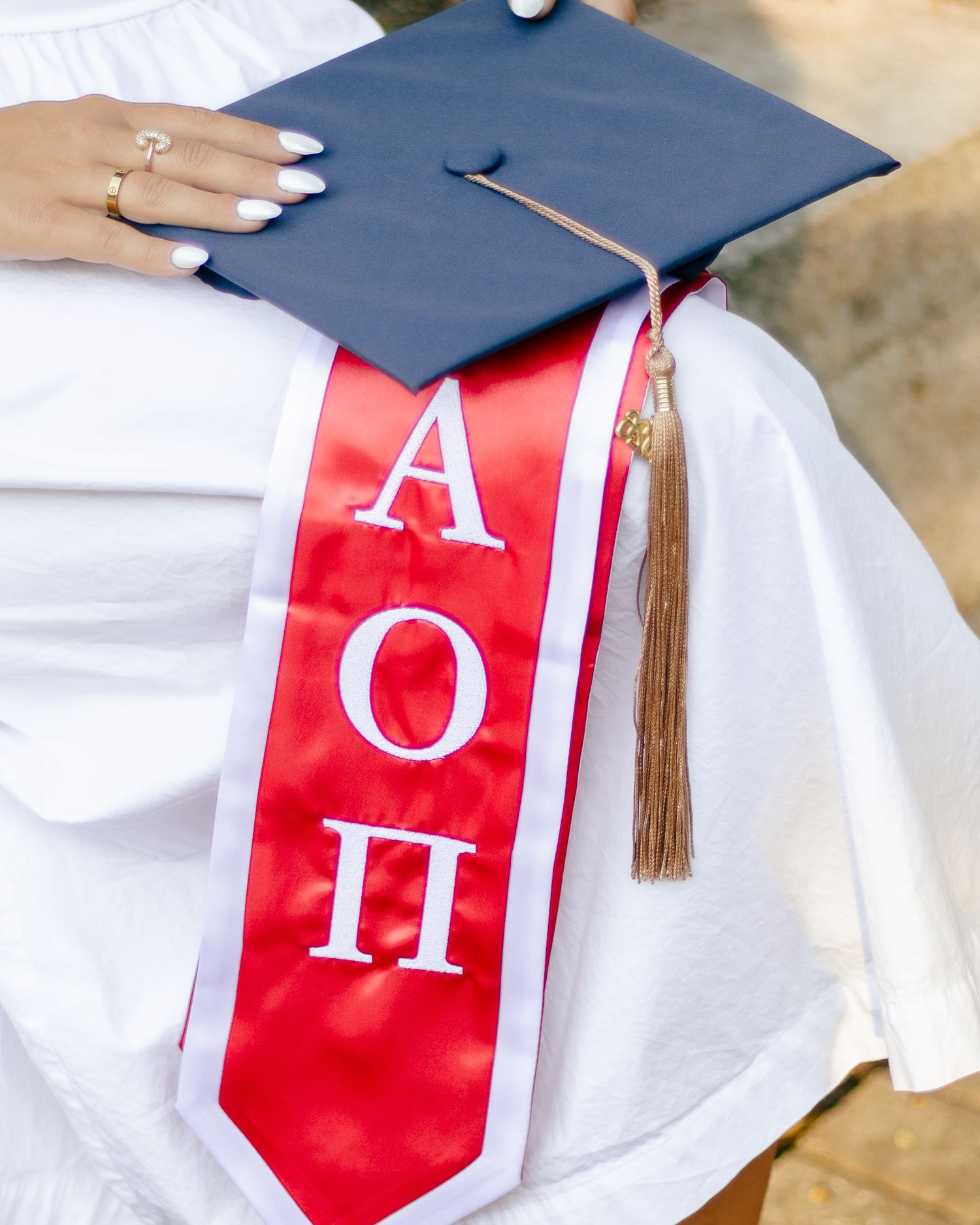 Graduate in a white dress holds a navy cap with tassel and a red AOII sorority graduation stole across her lap.
