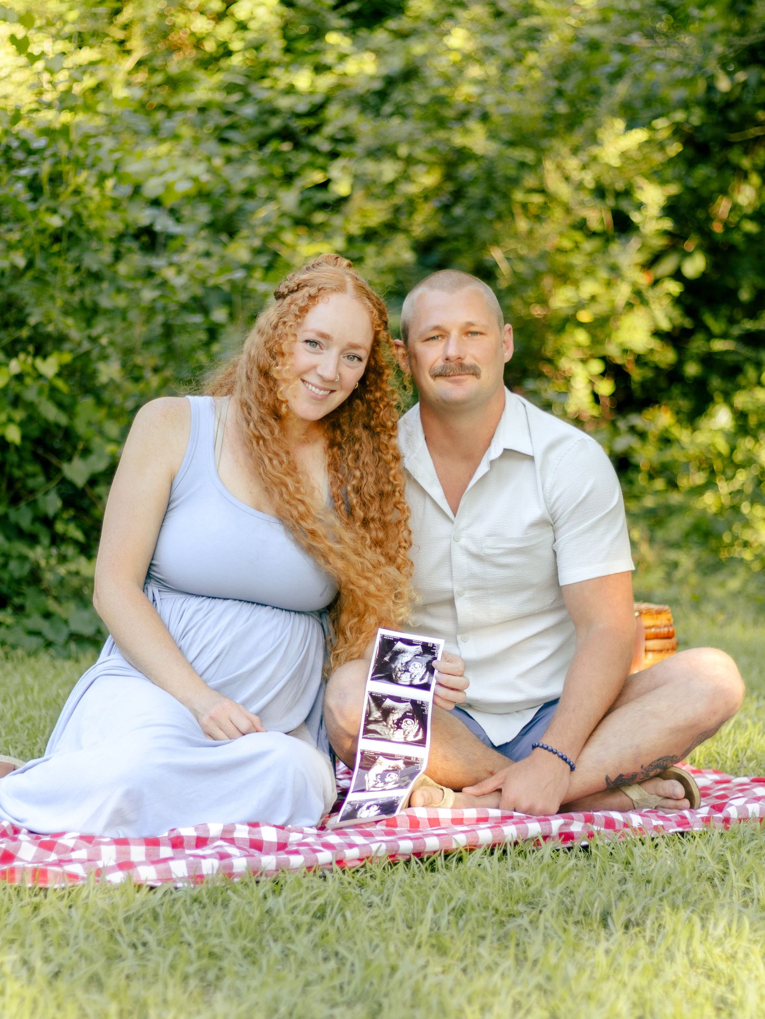 Smiling expectant couple sits on a red gingham picnic blanket in a sunny park, holding ultrasound photos of their baby.