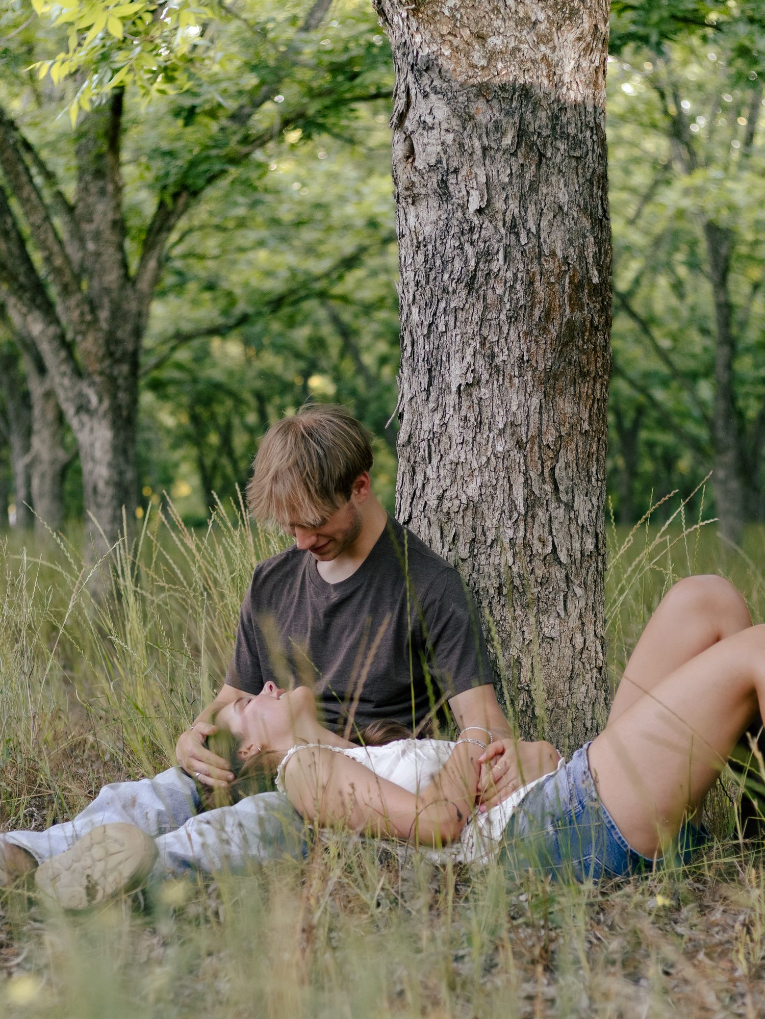 Couple relaxing in tall grass under a tree, a man leaning against the trunk as a woman rests her head in his lap in a sunlit forest.