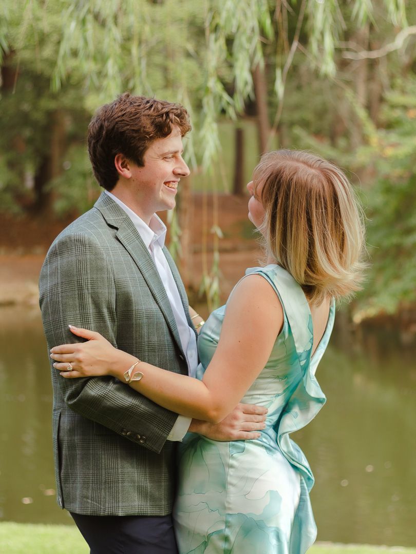 A happy man in a green plaid blazer smiles as a woman in a flowing aqua dress hugs him beside a peaceful pond surrounded by trees.