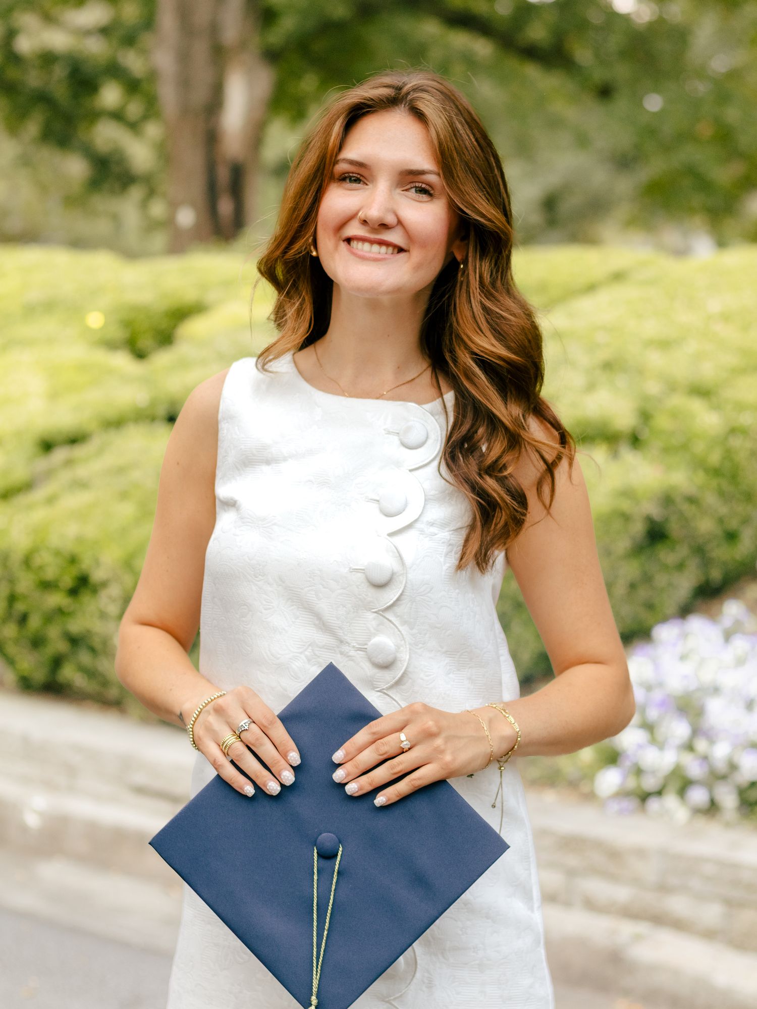 Smiling woman in a white sleeveless dress holds a navy blue graduation cap outdoors beside green hedges and purple flowers.