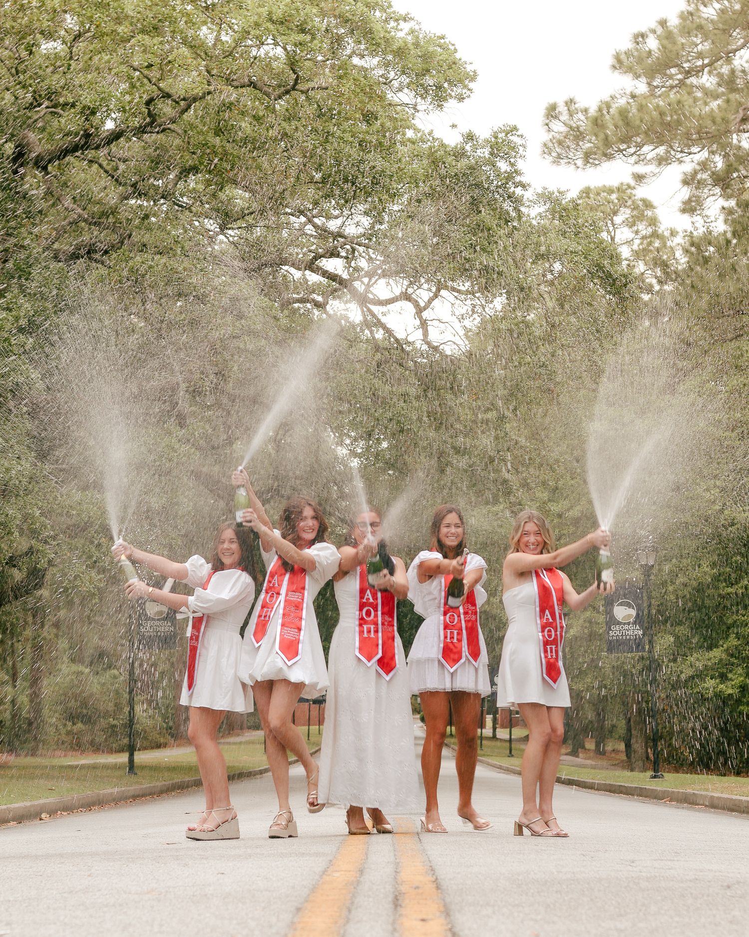 Five women in white dresses with red sashes spray champagne in celebration while standing on a tree-lined road.