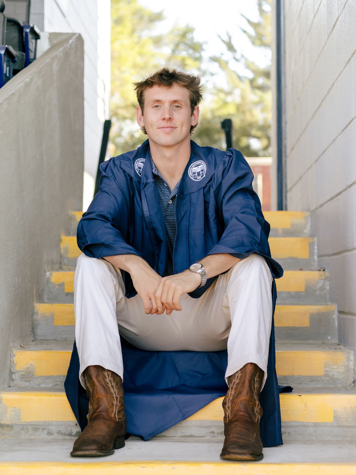 Young man in a blue graduation gown and brown cowboy boots sits smiling on yellow-edged concrete steps outdoors.