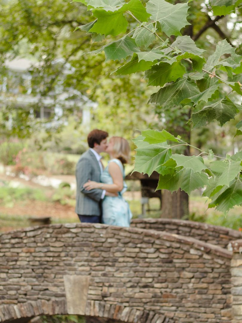 Couple in a gray suit and teal dress share a kiss near a stone arch bridge, framed by green leaves in a sunlit garden.