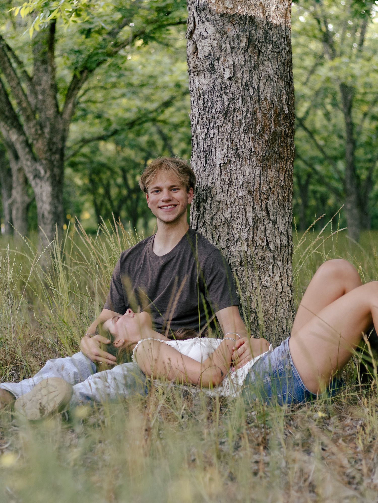 Smiling young man leans against a tree trunk in tall grass while another person rests their head in his lap in a sunny wooded park.