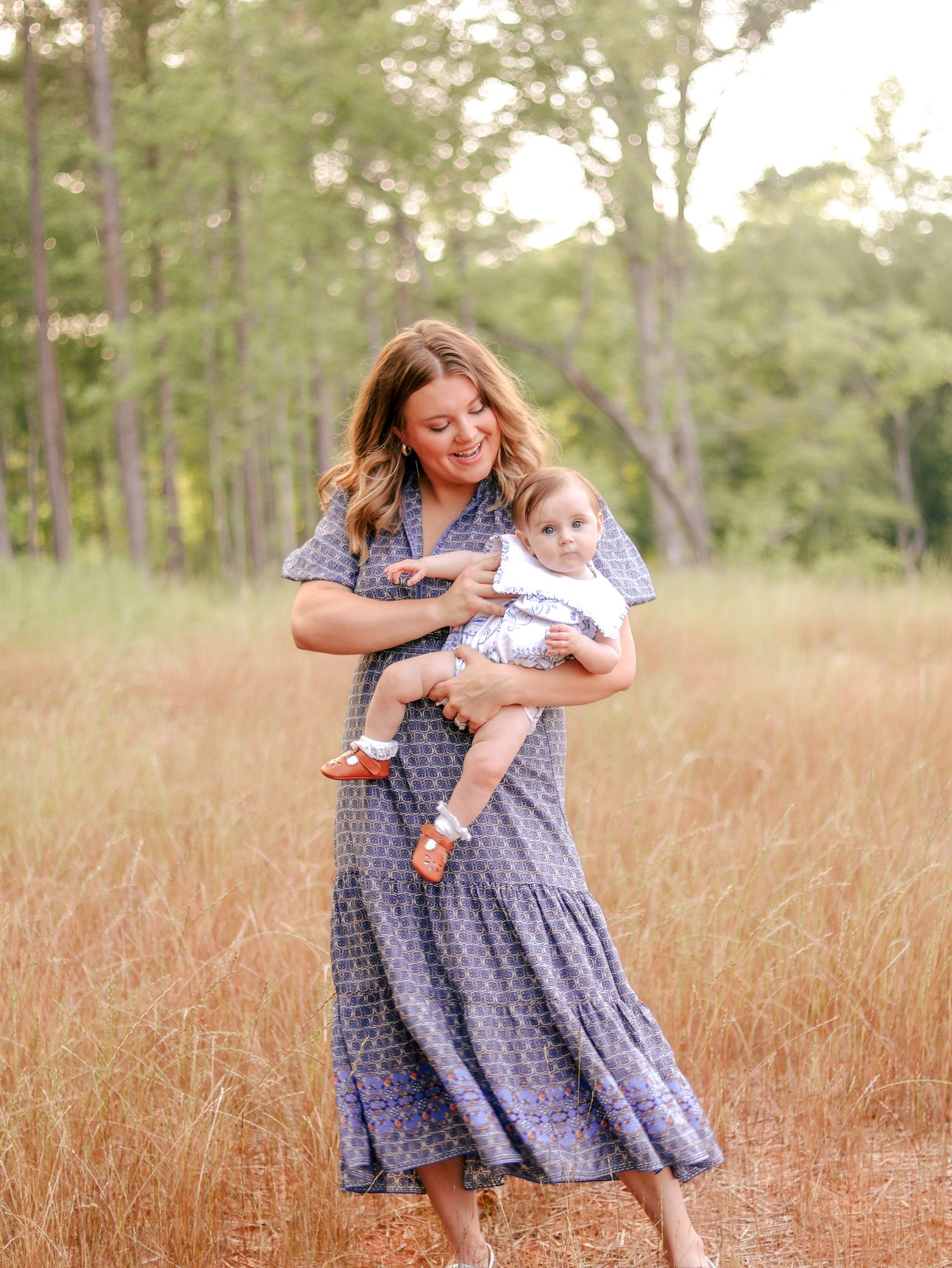 Smiling woman in a blue patterned dress holding a baby in orange shoes while standing in a golden grass field with tall trees behind.