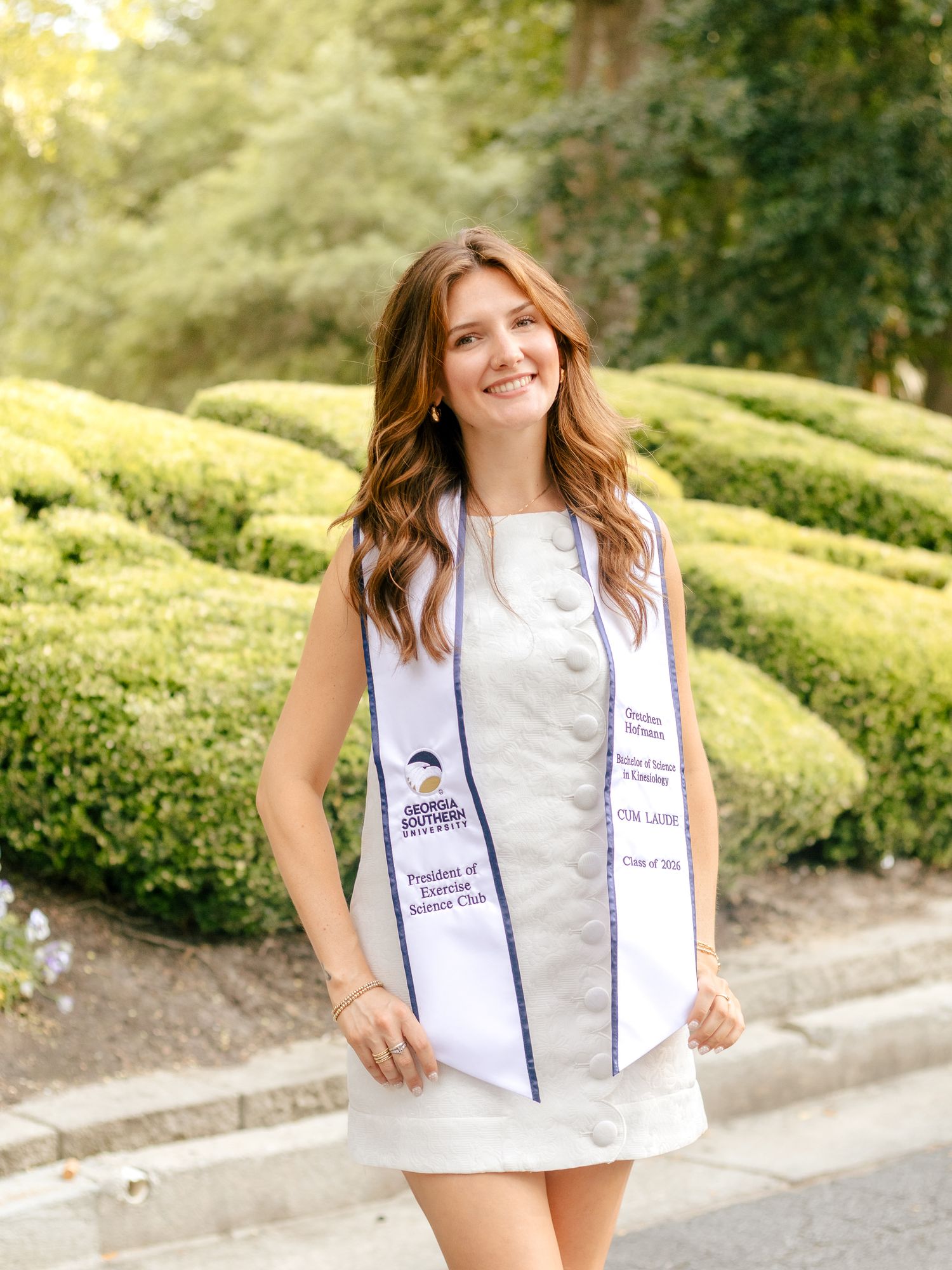 Smiling graduate in a white dress and Georgia Southern University graduation stole stands in front of trimmed green hedges outdoors.