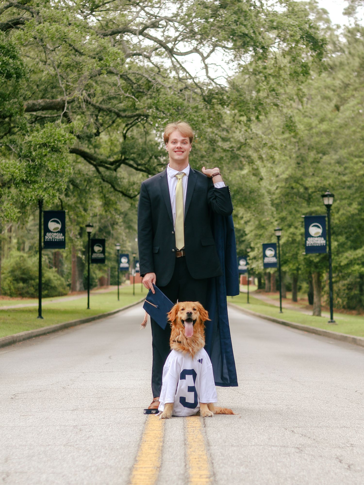 Young man in a suit holding a graduation gown stands on a tree-lined campus road beside a golden retriever wearing a number 3 jersey.