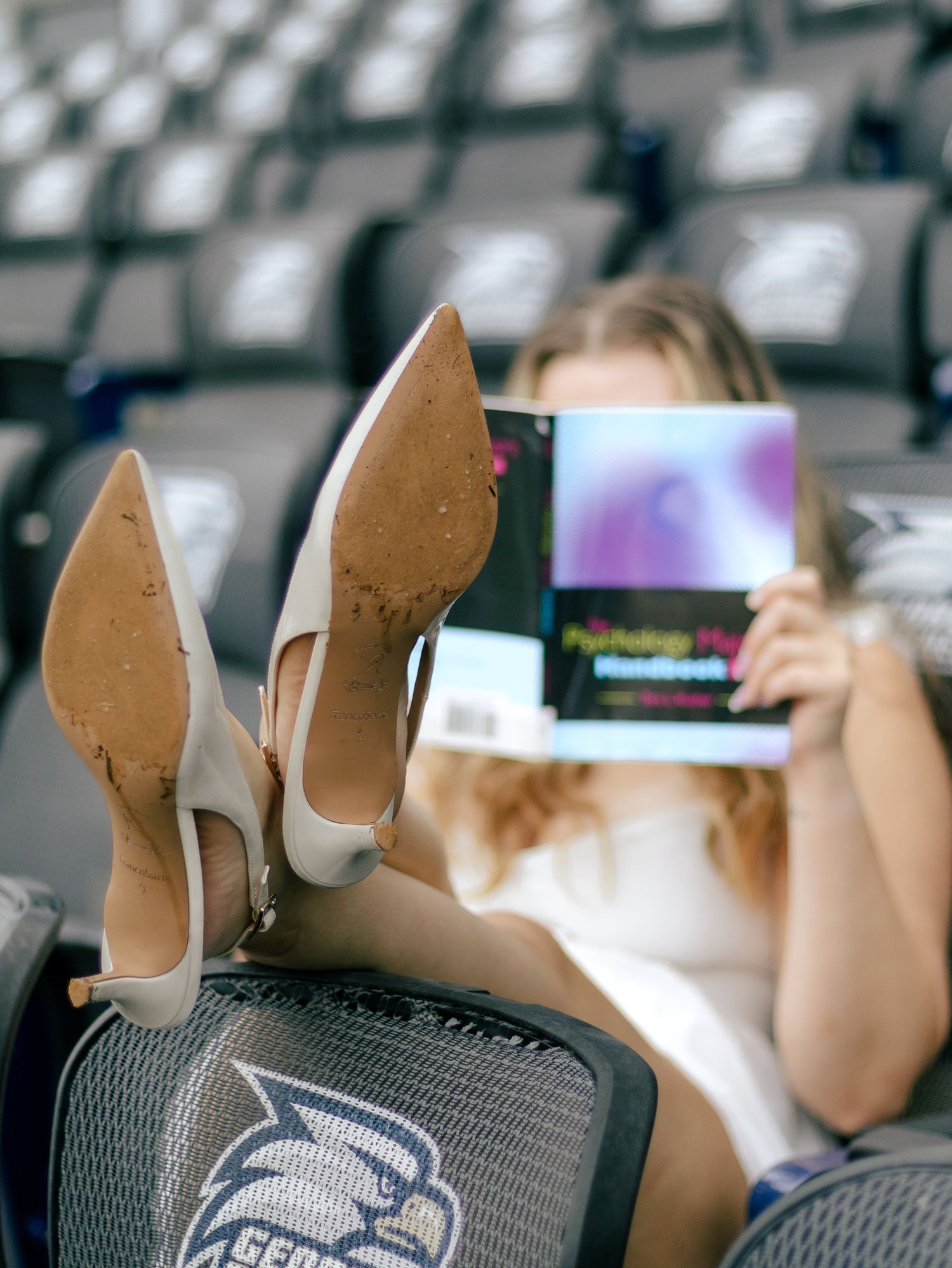 Woman in a white dress reads a psychology textbook in arena seats, her tan high heels propped up on a chair bearing a blue bird logo.