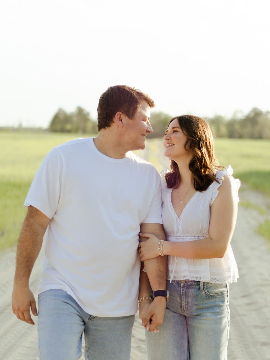 Smiling couple in white tops and jeans walk arm in arm down a sandy country path beside a green field at golden hour.
