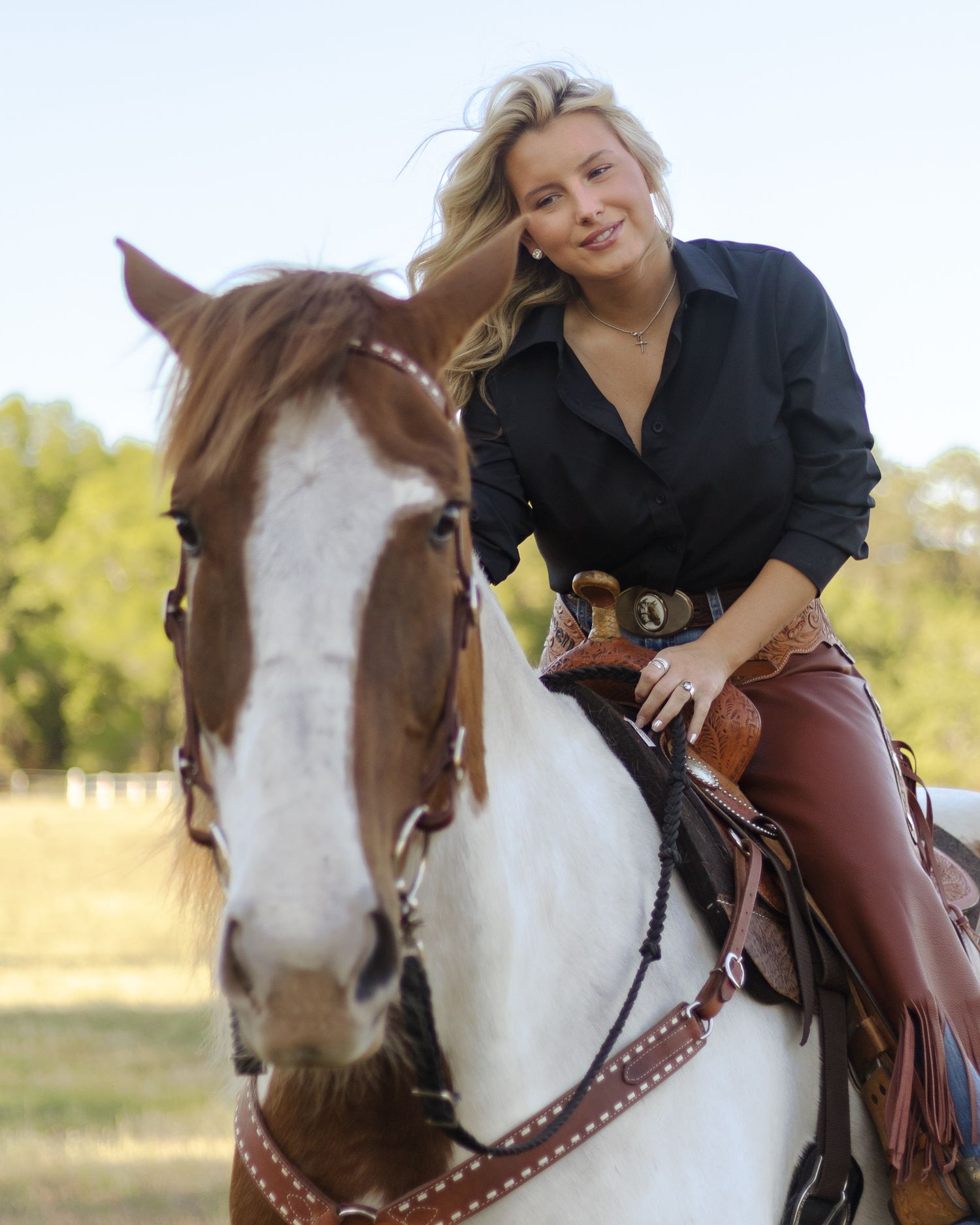 Smiling blonde woman in a black shirt and brown chaps rides a brown and white paint horse in a sunny grassy field.