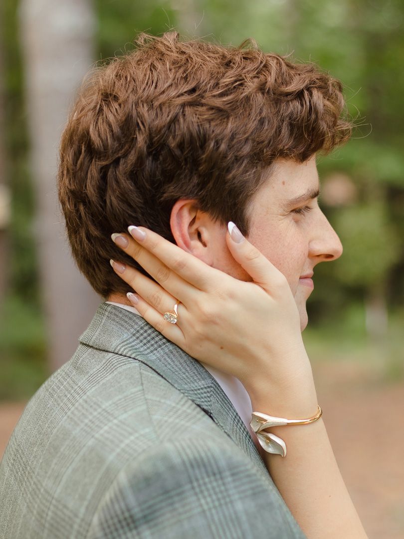 Close-up side profile of a man in a gray plaid blazer as a woman's hand with a ring and gold bracelet gently touches his cheek.