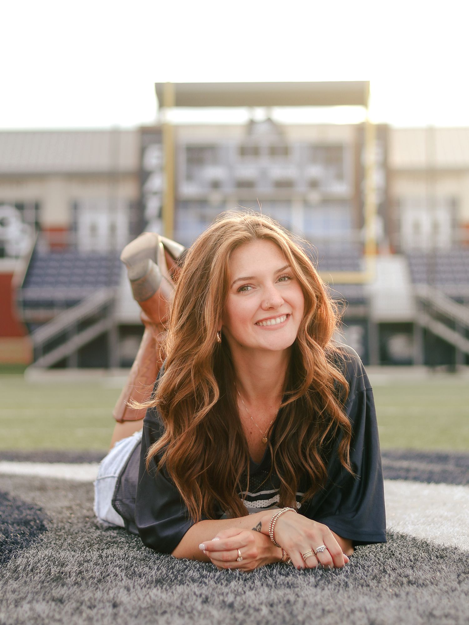 Smiling young woman with long wavy auburn hair lying on a turf football field with stadium bleachers softly blurred behind her.