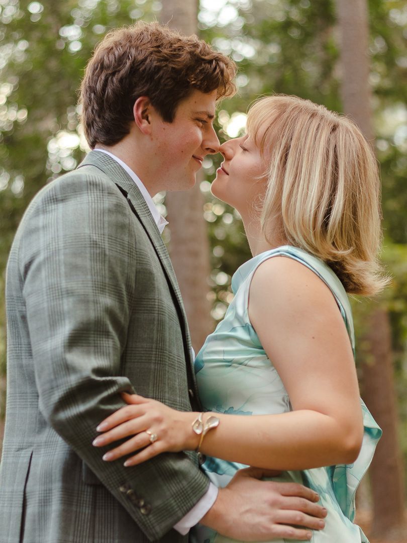 Close-up of a smiling couple nose to nose in a tender embrace, surrounded by softly blurred green trees at an outdoor engagement session.