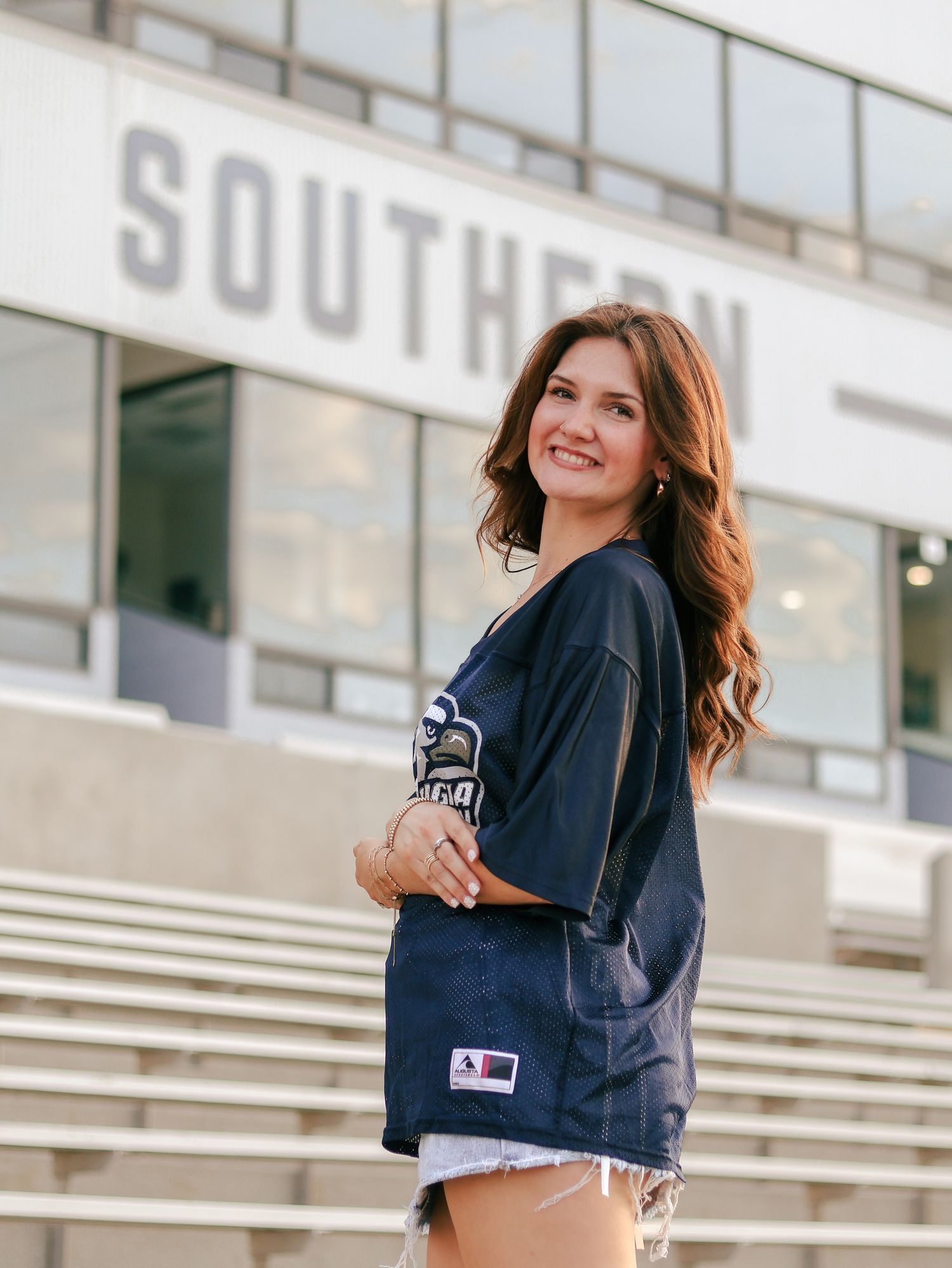 Smiling young woman with long wavy auburn hair wears a navy game day jersey and denim shorts while posing on empty stadium bleachers.