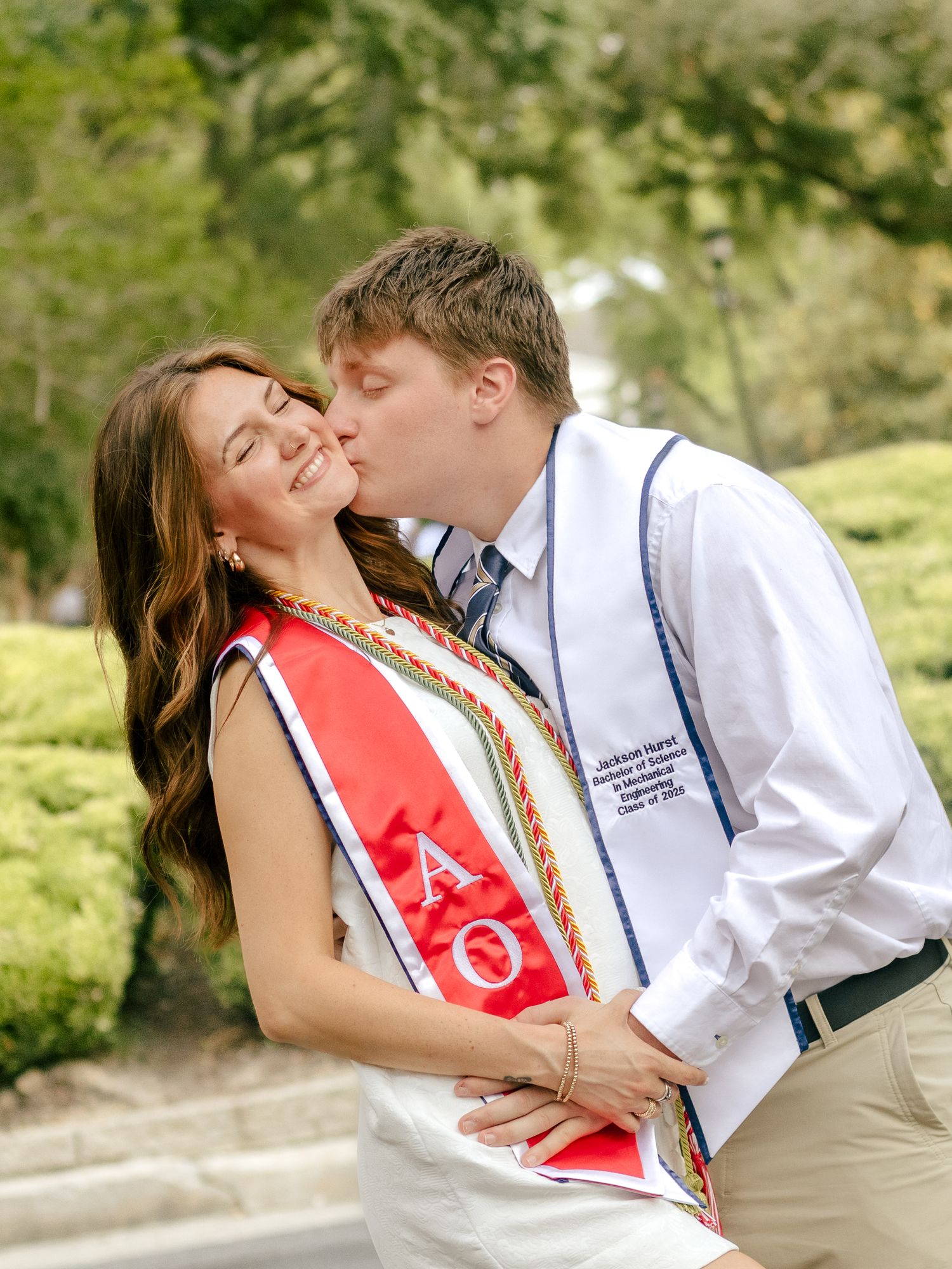 A young man kisses a smiling woman on the cheek while both wear graduation stoles outdoors in a leafy, green setting.