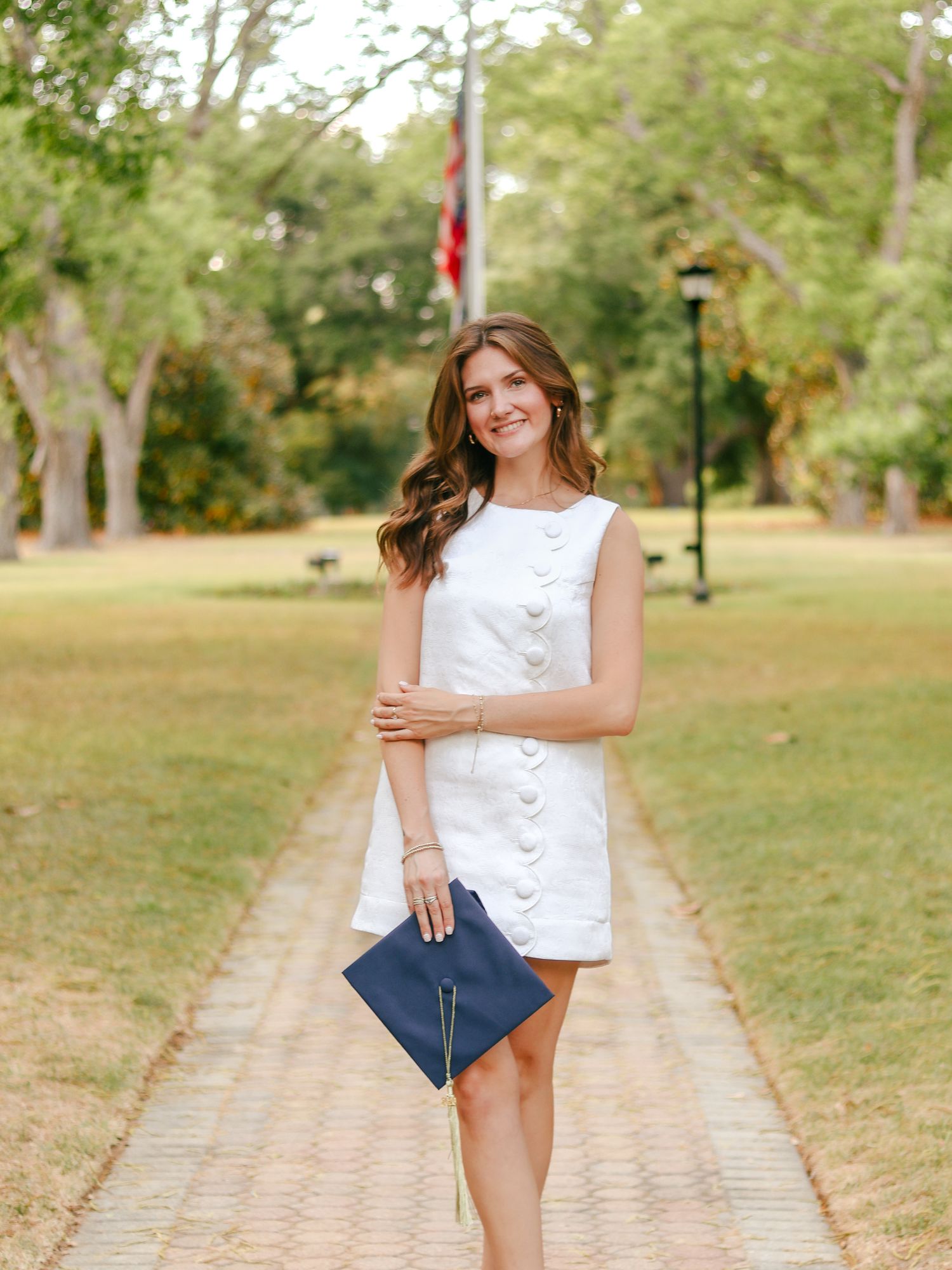 Woman in a white button-front dress stands on a brick path in a sunny park holding a navy graduation cap.