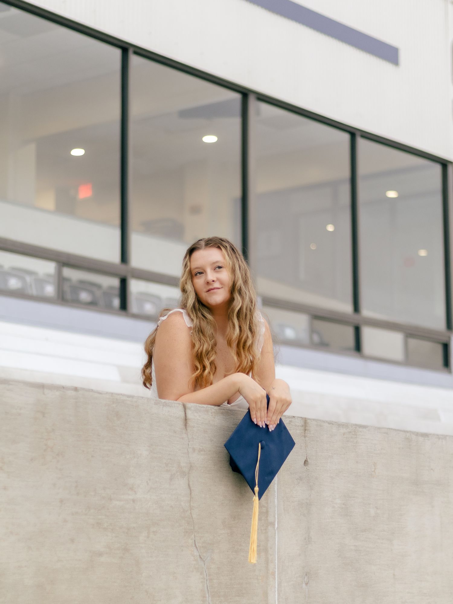 Smiling graduate with long wavy hair leans on a concrete ledge holding a navy blue graduation cap with a gold tassel.