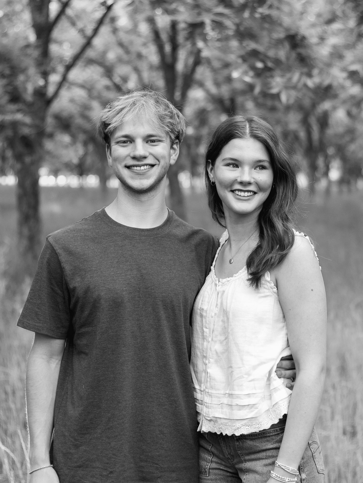 Black and white portrait of a smiling young man and young woman standing close together outdoors in a sunlit grassy park with trees.