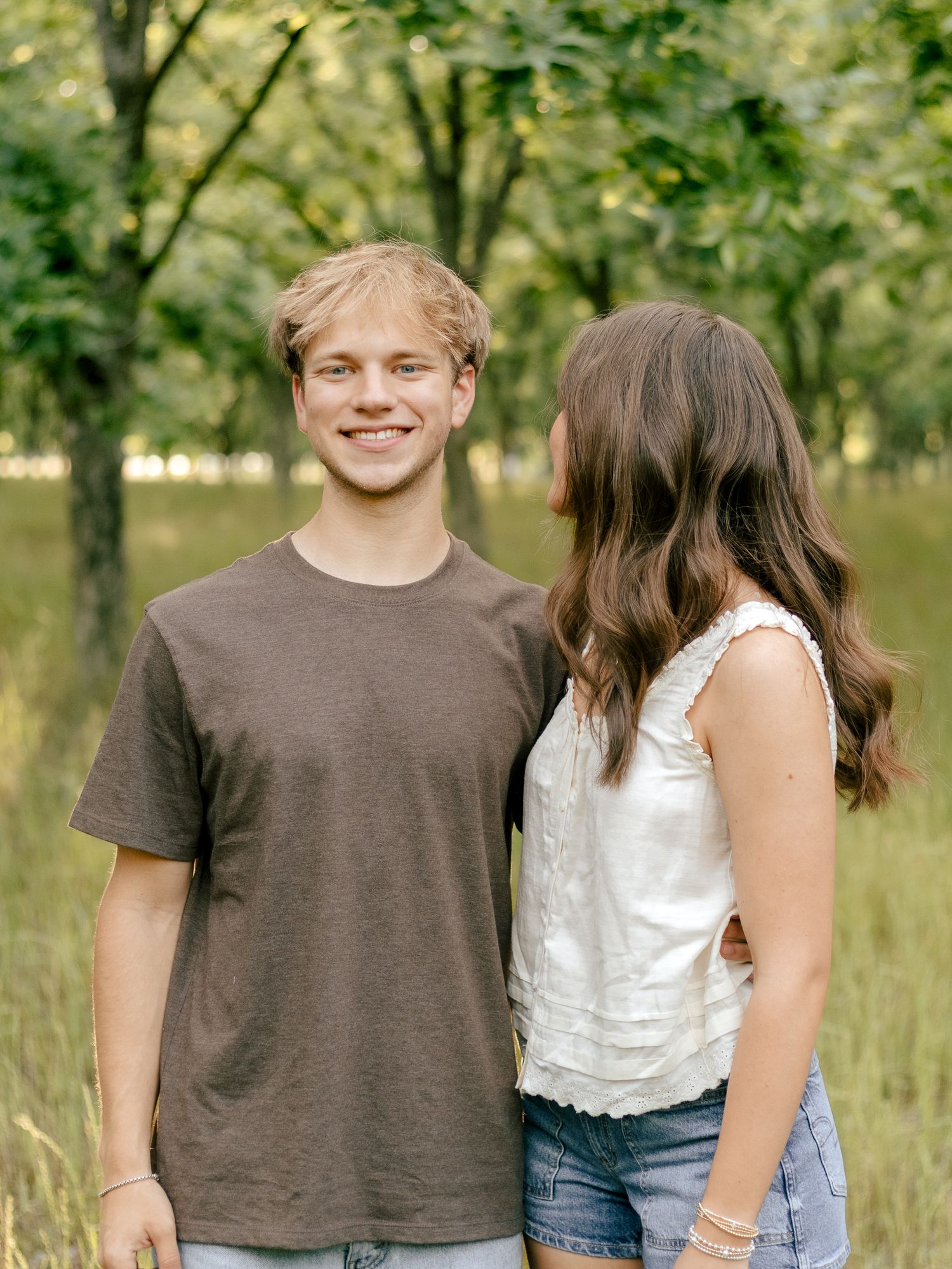 Smiling young man in a brown t-shirt stands beside a woman in a white top and denim shorts in a sunny, grassy orchard.