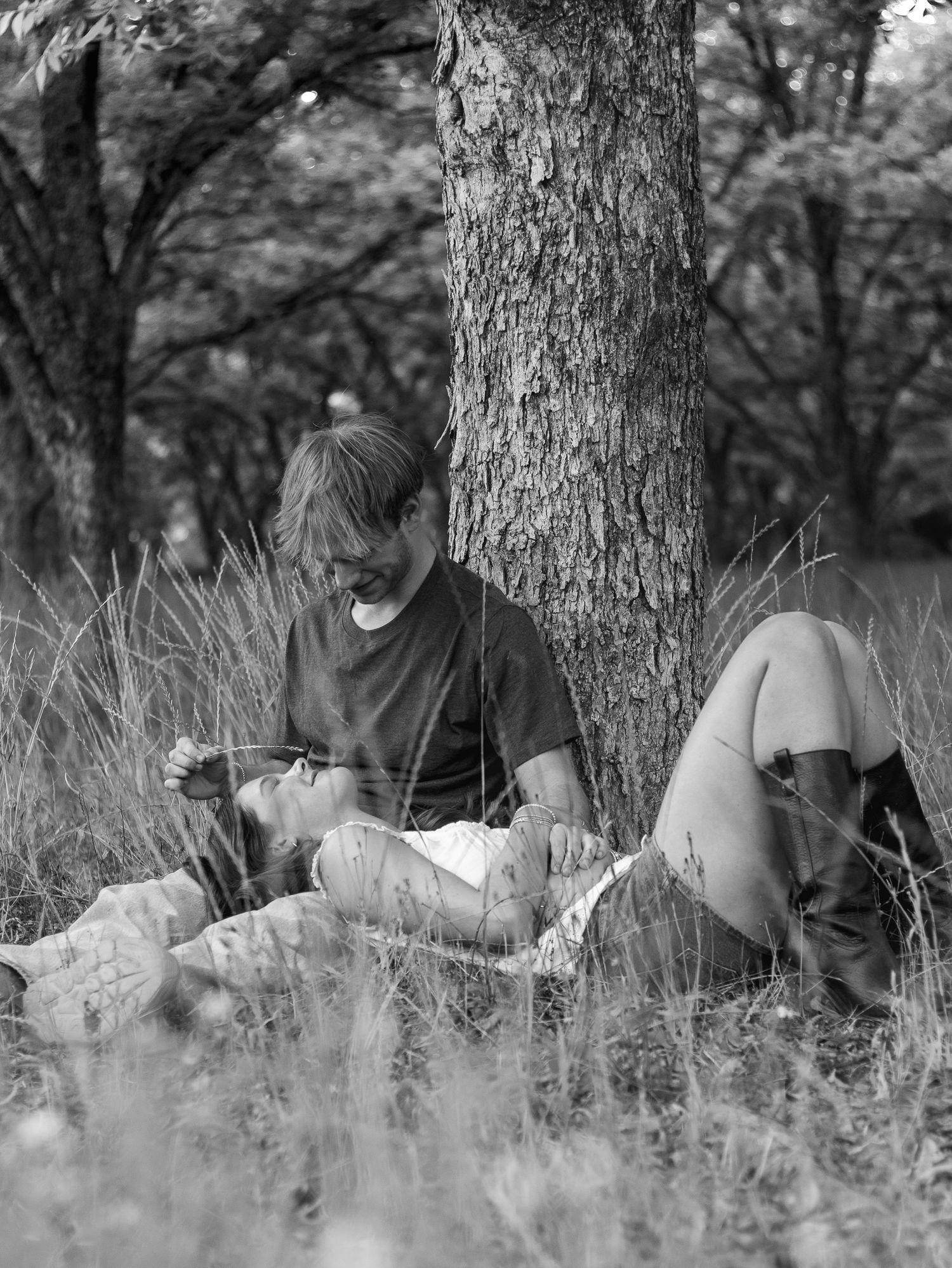 Black and white photo of a smiling man leaning against a tree trunk in tall grass while a woman in boots reclines beside him.