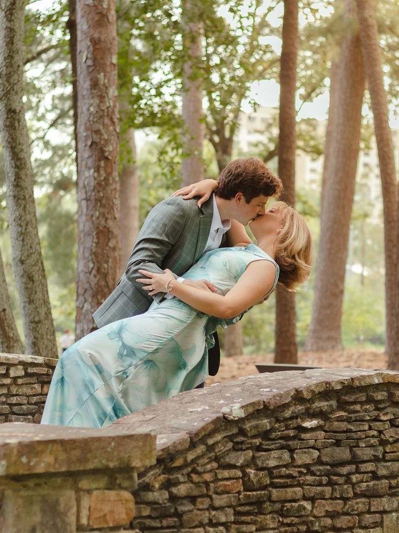 A man in a gray blazer dips a woman in a teal dress for a romantic kiss beside a stone wall in a sunlit wooded park.