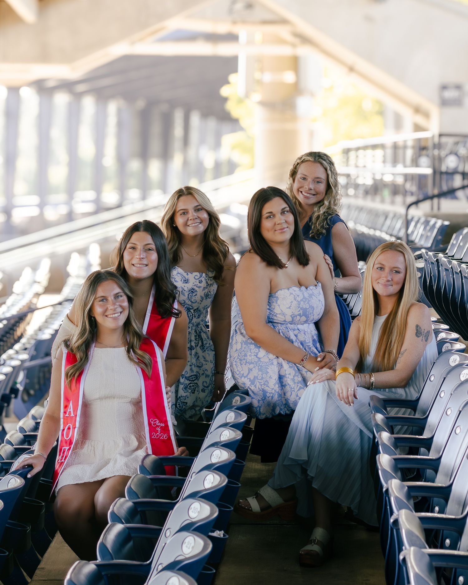 Five smiling young women in summer dresses pose together on blue stadium seats, two wearing red graduation sashes.
