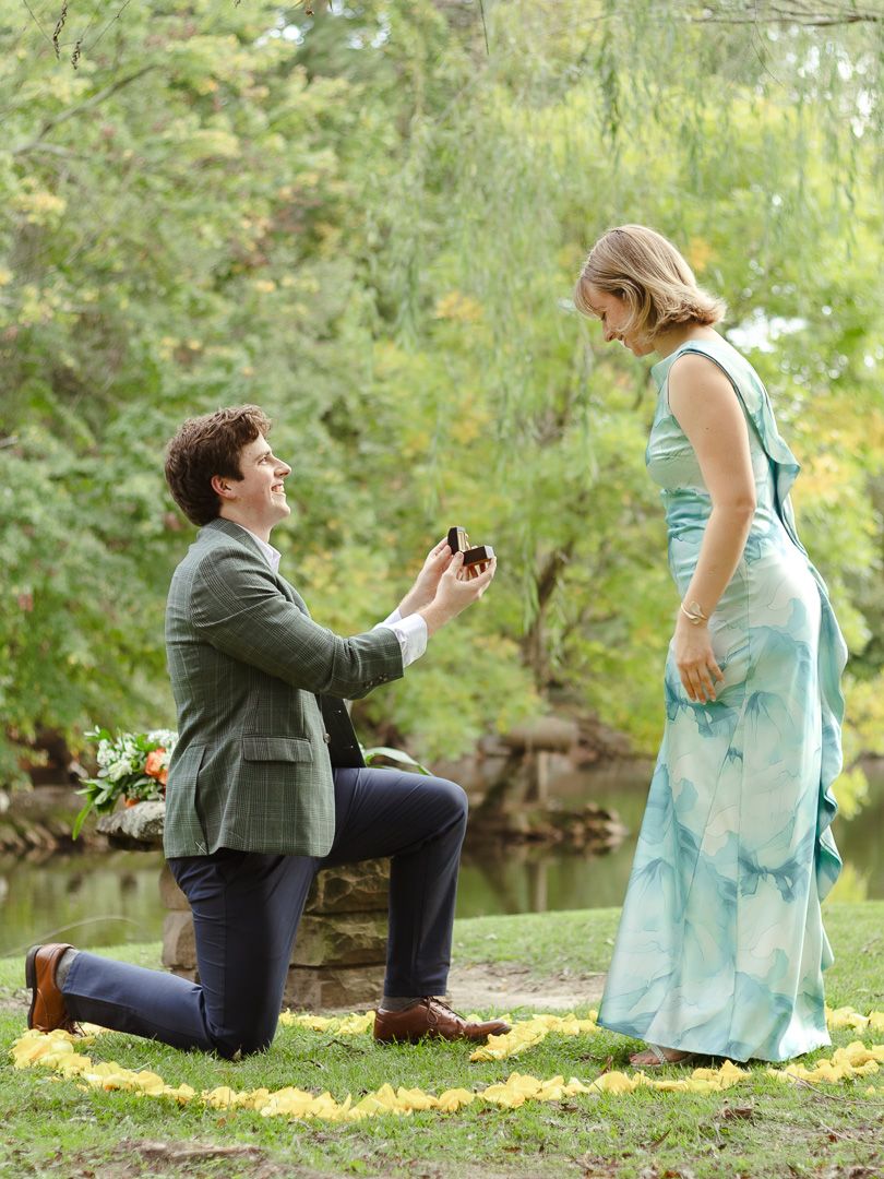 A man in a tweed blazer kneels on the grass holding a ring box while proposing to a smiling woman in a blue dress by a lake.
